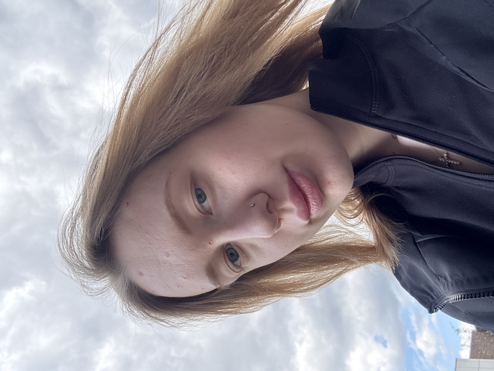 An eye-level shot captures a young individual, likely female, with light brown hair, looking directly at the camera. She has blue-gray eyes and a small, silver stud piercing on her nose. Her expression is neutral. She is wearing a black zippered jacket, partially unzipped to reveal a silver-colored cross pendant on a necklace around her neck.
The background consists primarily of an overcast or partly cloudy sky with visible white and gray clouds. A faint outline of a building or structure is visible in the bottom right corner. The image is taken from a low angle, looking up at the subject, which makes the sky a prominent feature. The overall scene suggests an outdoor setting during the daytime in Rostov-on-Don, Russia. The image is rotated, giving the impression that the subject's head is significantly tilted relative to the horizon. No text is visible in the media.