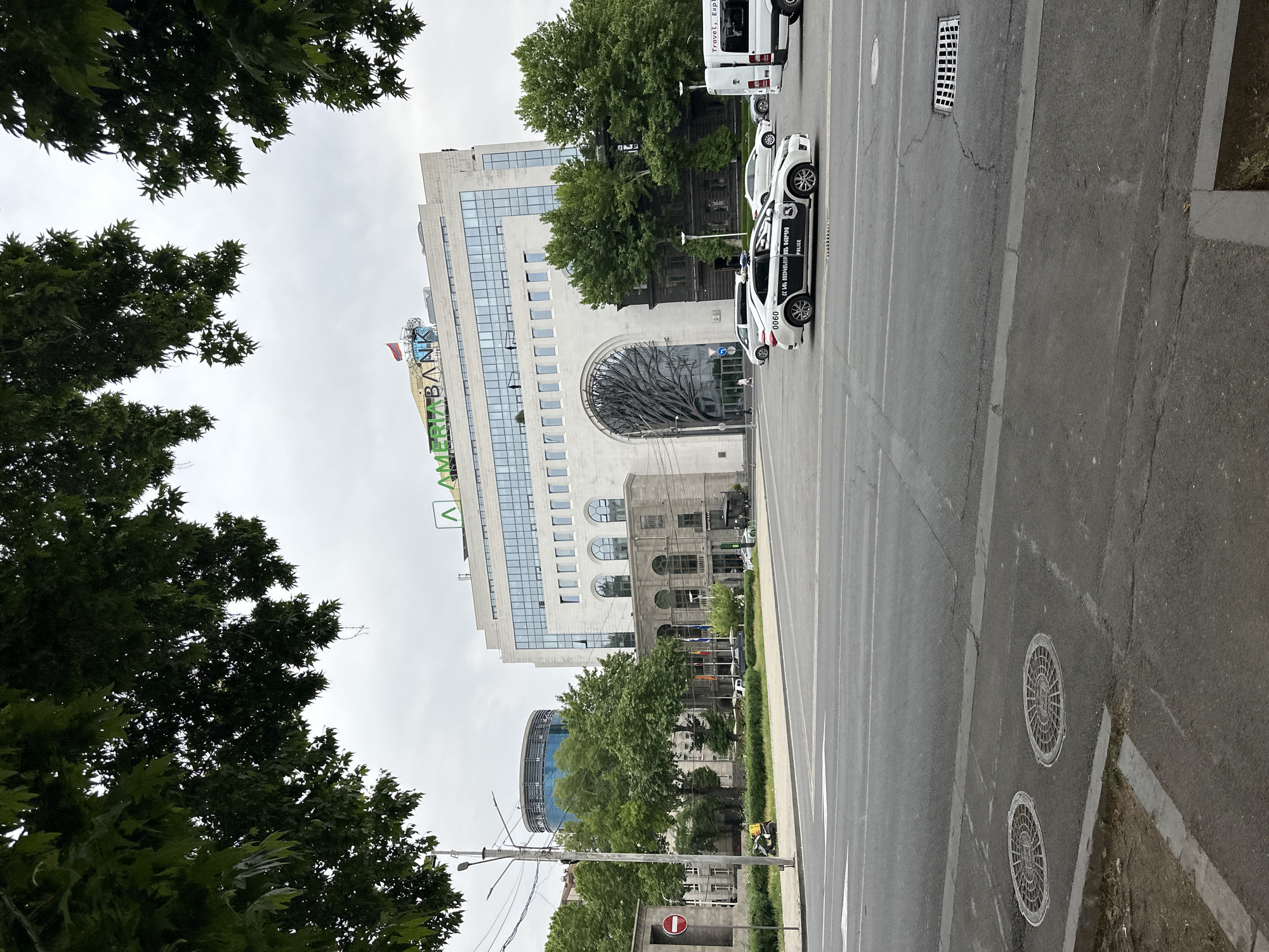 The image shows a street view in Yerevan, Armenia. In the background, a modern, multi-story building with the "AMERIABANK" logo is visible, indicating the presence of the bank's headquarters or a branch. Adjacent to it is a building with a large, arched entrance featuring a decorative metal tree-like structure. Several vehicles, including police cars and a white van, are parked on the side of the road. The street is lined with trees, partially obscuring other buildings in the distance, including a cylindrical glass structure. The road surface is paved with visible lane markings and two round manhole covers on the right side. Streetlights and utility poles are also visible, along with some traffic signage.