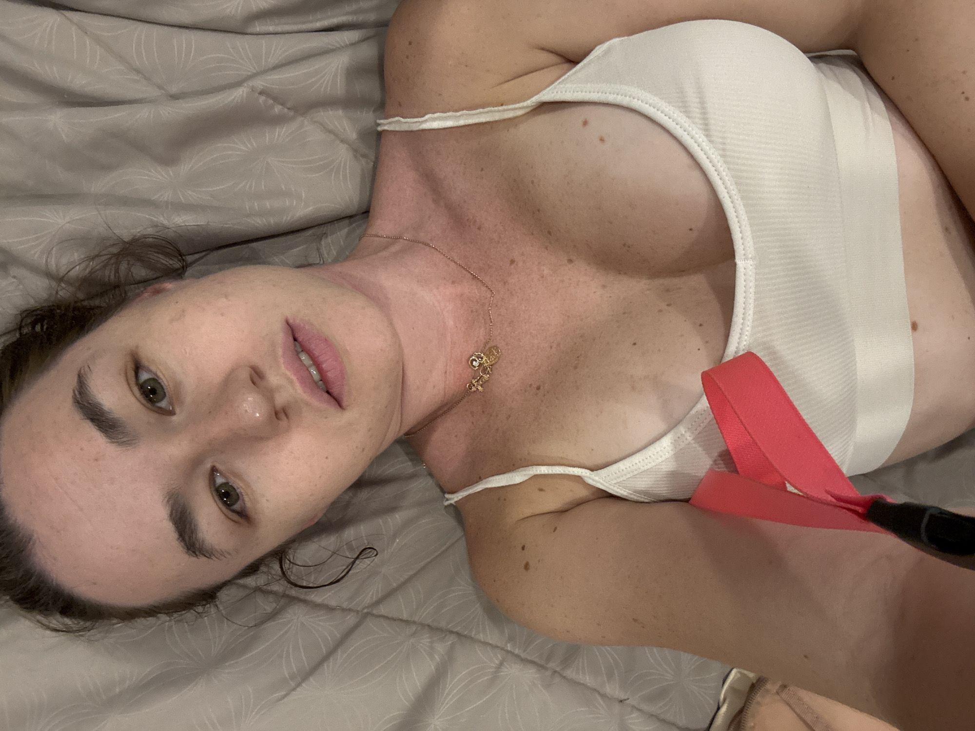A young woman with light brown hair and green eyes is lying down on a grey, textured blanket. She is wearing a white ribbed tank top. A thin gold necklace with a small charm hangs around her neck. Her skin is light and appears to have freckles. A pink strap is visible, held in her right hand, which is partially obscured by a black object. Her body is positioned diagonally across the frame, with her head on the left and her torso extending to the right. The lighting is dim and appears to be indoor ambient light. The location is identified as Rawai, Thailand.