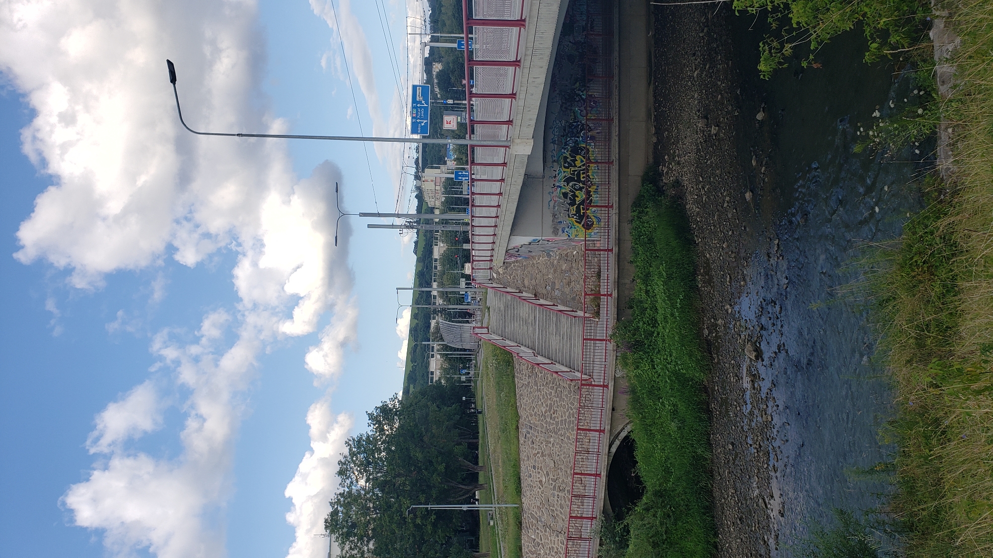 The image shows a river flowing under a bridge with a pedestrian walkway. The bridge is made of concrete and has red railings. There is graffiti on the underside of the bridge. The riverbank is covered in green grass and rocks. In the background, there are trees and buildings. The sky is blue with some white clouds. The scene appears to be located in the city of Prešov, Slovakia, as indicated by a blue road sign that reads "Prešov."