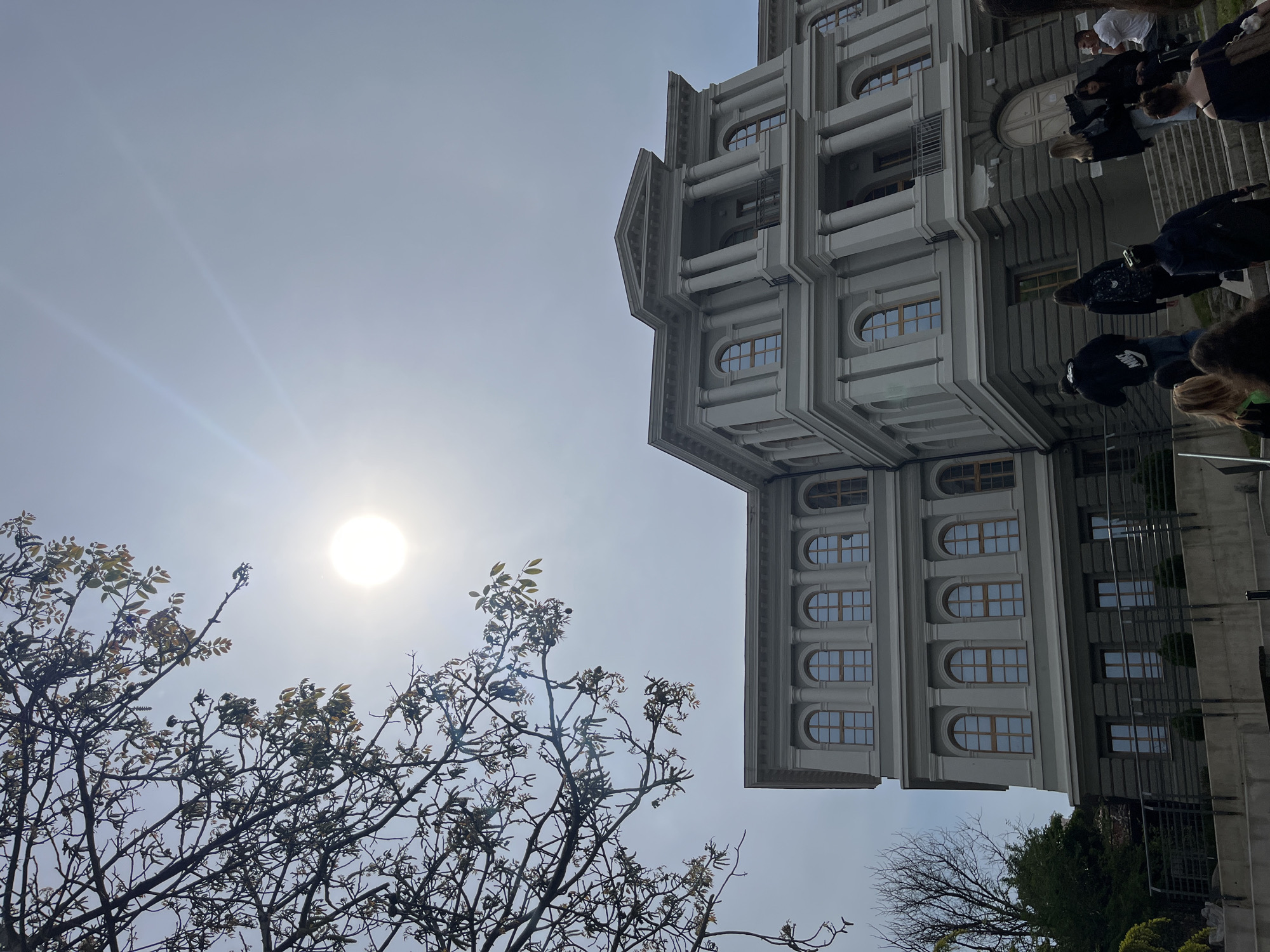 The image captures a building with a distinctive architectural style, potentially located in Plovdiv, Bulgaria. The building is characterized by its multiple stories and repetitive window patterns, appearing in a light grey or white color. To the left of the building, tree branches are visible, partially obscuring the sun, which shines brightly in the sky. The photo appears to have been taken from a low angle, looking upwards towards the building and sky. In the original image, some people are visible, but they are blurred.