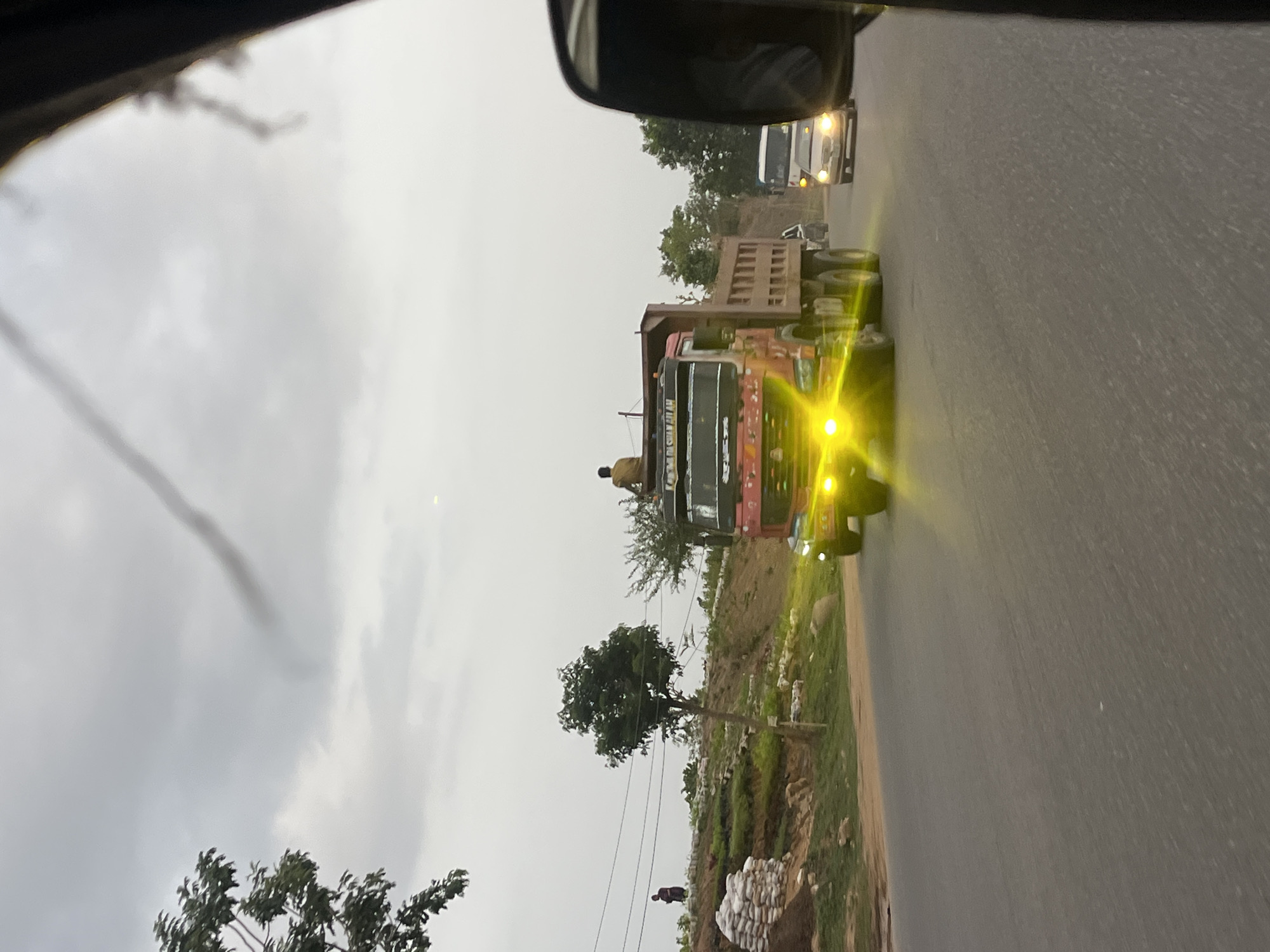 The image shows a road in Karmajiji, Nigeria. A large, red and black truck with bright yellow headlights dominates the scene, traveling on the road. A man is sitting on the truck's roof. In the background, another white vehicle is visible. Alongside the road, there is some vegetation and a pile of white bags or sacks. A person appears to be near the pile. The sky is overcast, indicating a cloudy day. The view is taken from inside a vehicle, showing part of the car's interior and side mirror.