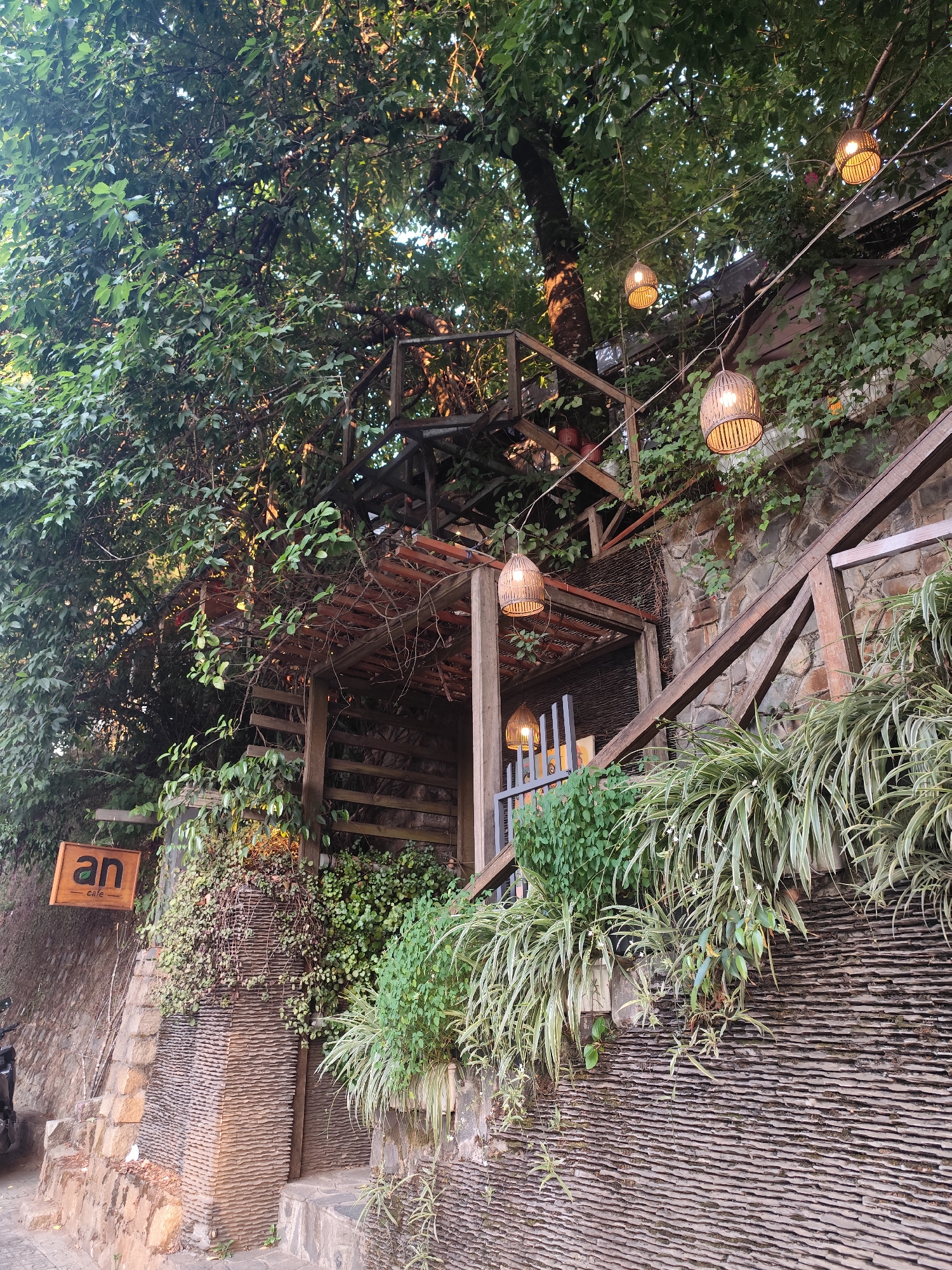 An exterior shot shows a cafe in Da Lat, Vietnam, overgrown with greenery. The architecture consists of wood and stone elements. String lights hang from the trees. A sign reads "an cafe". Plants grow out of the stone wall. A motorcycle is partially visible on the bottom left.