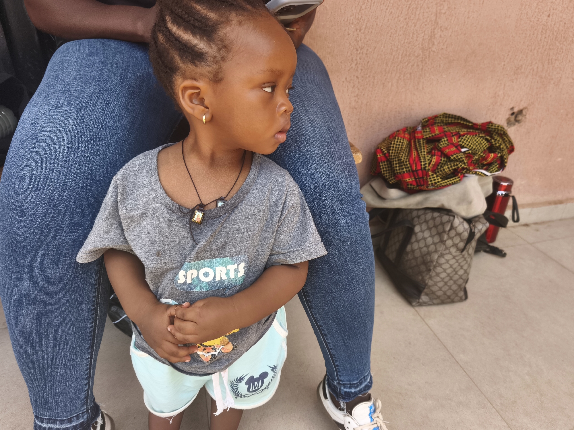 A young child stands in front of a person sitting down, presumably on the tile floor of a building in Jos, Nigeria. The child has dark skin, braided hair, and is wearing a gray shirt with "SPORTS" printed on it, along with light blue shorts with a cartoon graphic, a gold earring, and a necklace with two pendants. The person behind the child is wearing blue jeans and sneakers. To the right of the child is a pile of folded fabric including red and yellow patterned cloth and a red insulated drink container is nearby.