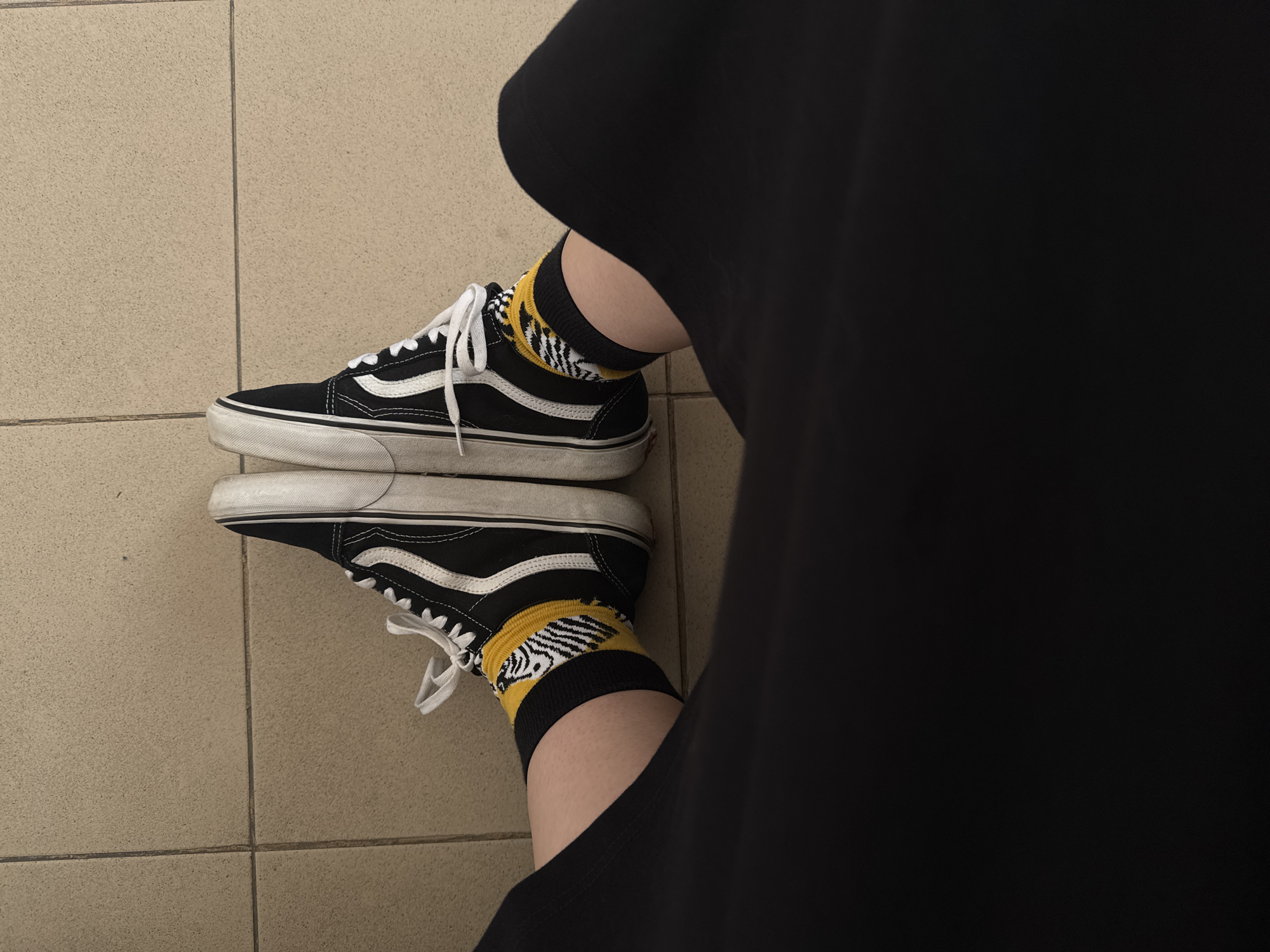 The image shows a person's legs and feet wearing black Vans Old Skool sneakers and black socks with a yellow and white zebra pattern. The person is wearing black pants. The feet are positioned in a V-shape on a tiled floor. The tiles are rectangular and beige with visible grout lines. The background is out of focus. The image does not contain any discernible features or context that would allow for its location to be identified as Kaliningrad, Russia.