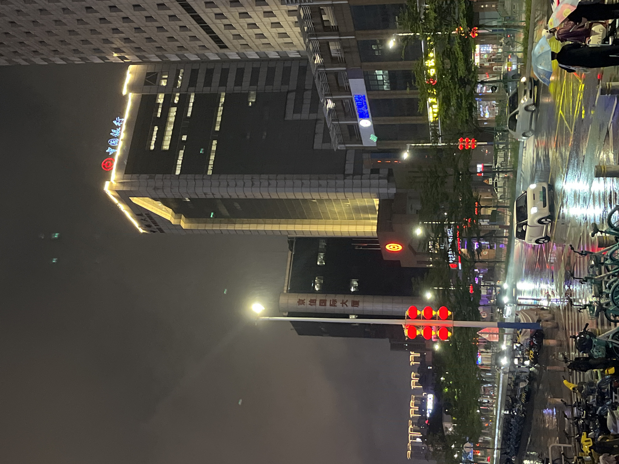 The image captures a night scene in the city of Nanhai District, China, under rainy conditions. A tall, modern building with illuminated signage reading "中国银行" (Bank of China) dominates the skyline. To its left, another tall building displays vertical text. In the foreground, a wet street reflects the city lights and headlights of moving vehicles. A traffic light with red signals is prominent. Rows of parked bicycles and scooters line the sidewalk. Several people are present, some holding umbrellas, suggesting they are navigating the wet weather. The overall atmosphere is urban and atmospheric, with the rain creating a reflective and somewhat subdued ambiance.