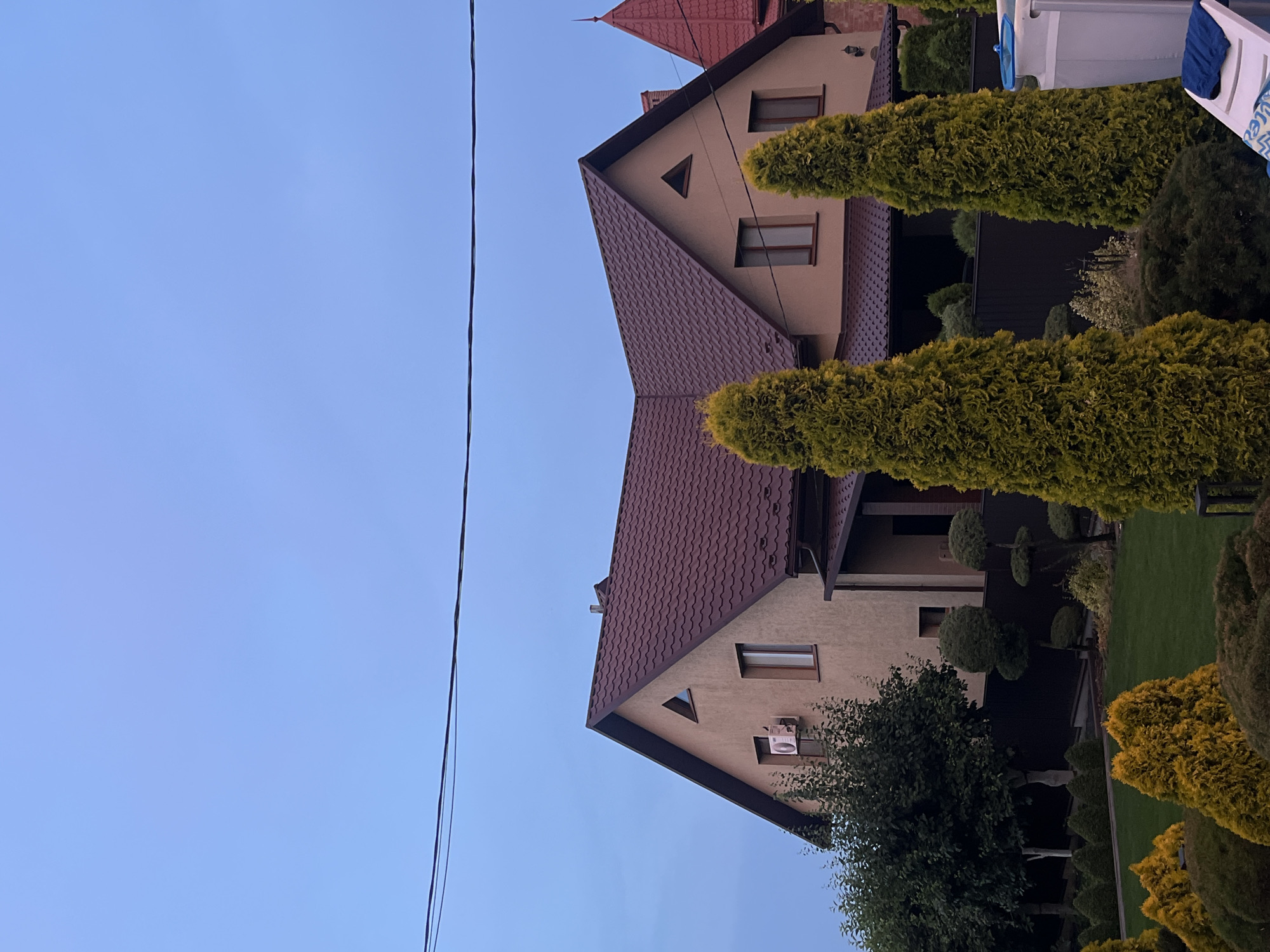 The image shows a residential building in Koroviia, Ukraine, against a clear blue sky. The house is a two-story structure with a brown, tiled roof and beige exterior walls. Several windows are visible, some with dark frames. An air conditioning unit is mounted on the exterior wall of the upper floor. In the foreground, there is a well-maintained garden with manicured trees and shrubs, including several tall, cone-shaped evergreen trees and smaller, yellow-leafed bushes. A dark fence separates the garden from the house. To the right of the frame, a portion of a white structure, possibly a pool or deck, is visible. The overall scene depicts a suburban or rural residential property.