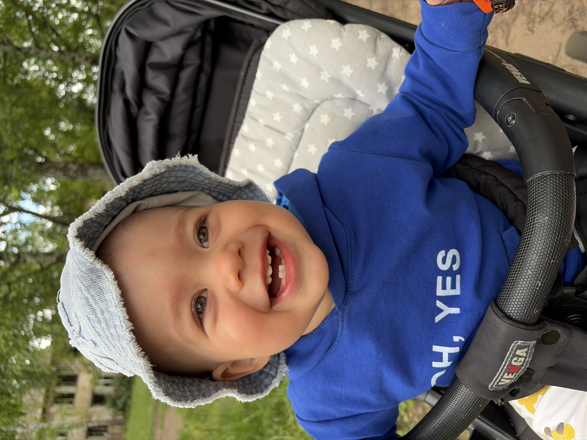A close-up, eye-level shot captures a smiling baby sitting in a stroller outdoors. The baby, wearing a light blue denim bucket hat and a bright blue hooded sweatshirt with white text that reads "OH, YES," looks directly at the camera. The stroller has a black frame and a gray padded insert with white star patterns. In the background, trees with green foliage and a weathered, multi-story building are visible, suggesting a location in an urban or suburban area, possibly Chaikovsky, Russia, given the context. The lighting is natural daylight, and the focus is on the baby's face, highlighting its joyful expression and the details of its clothing and the stroller.