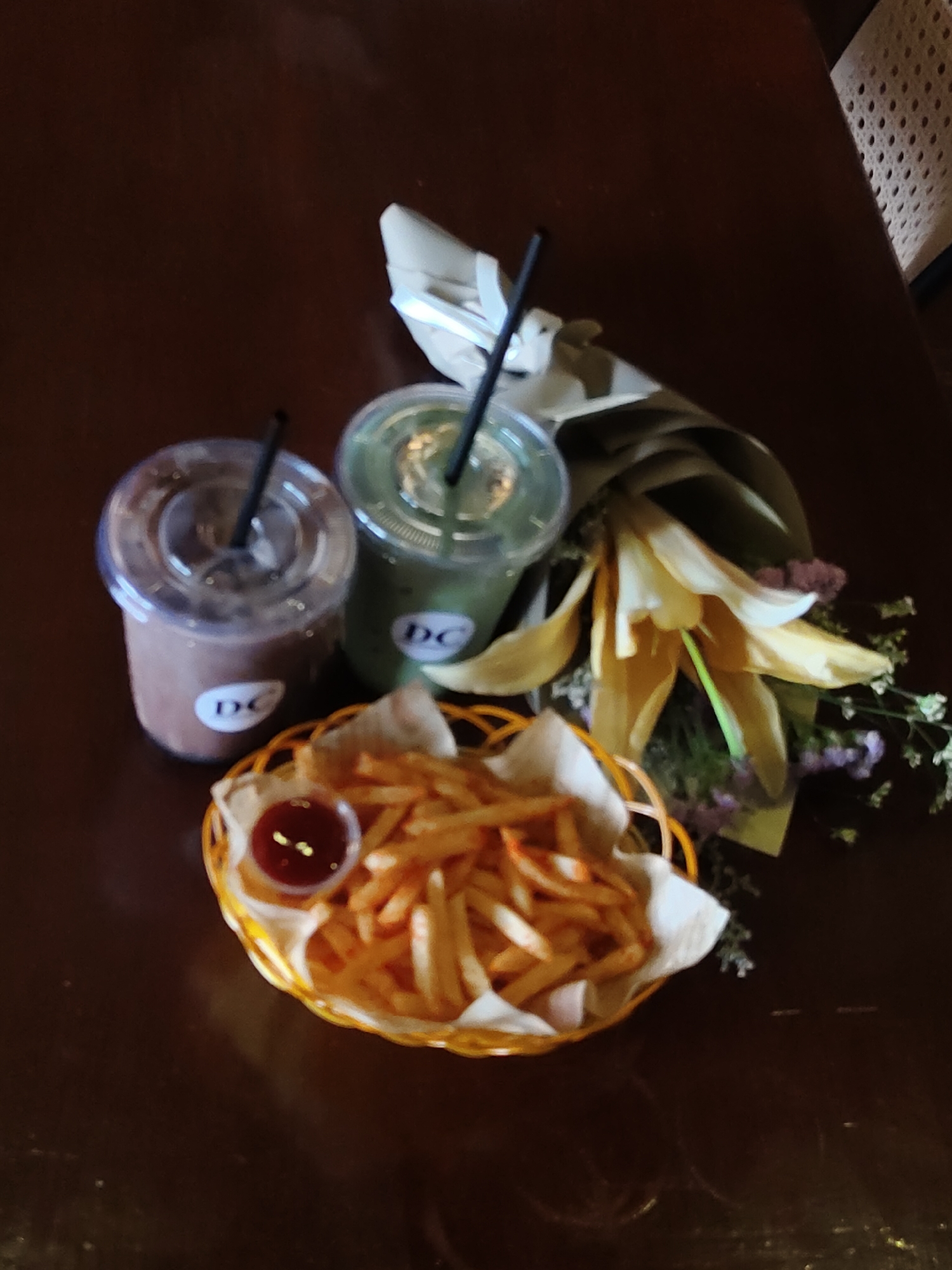 The image displays a dark brown wooden table with a basket of french fries and ketchup in a small dipping container. Beside the basket are two clear plastic cups with black straws, one containing a brown beverage and the other a green beverage. A bouquet of flowers, predominantly yellow lilies with some smaller purple flowers and greenery, is positioned behind the drinks. The logo "DC" is visible on both cups. The overall lighting suggests an indoor setting, possibly a cafe or restaurant. No people are visible in the image. The location is identified as Davao City, Philippines, though there are no visual cues in the image to confirm this specific location.