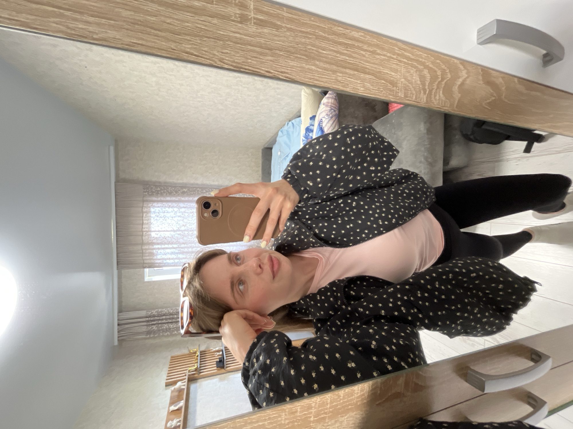 A woman is seen in a mirror selfie. She is wearing a pink top and a black jacket with a white floral pattern, and black leggings. She is holding a brown smartphone in her right hand. A pair of sunglasses is on the left side of her head. The mirror shows a bedroom interior with a bed, blinds on a window, and a dresser. The location is likely Chaikovsky, Russia.