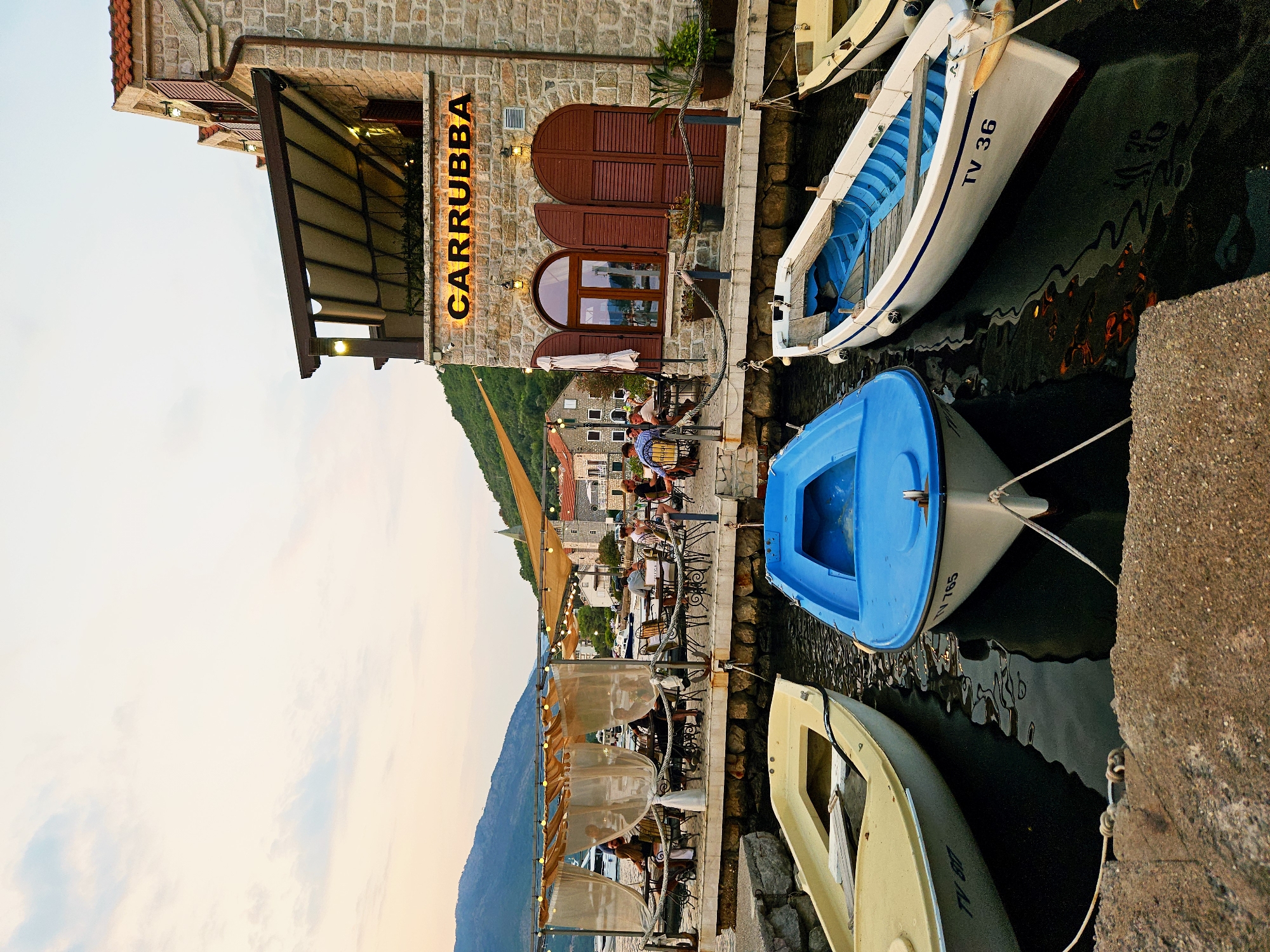 This is a photograph of a waterfront restaurant in Tivat, Montenegro. The restaurant, named "CARRUBBA," is a stone building with a covered outdoor seating area. Several people are dining at tables under the covered area. In the foreground, three small boats are docked in the water: a yellow boat, a blue boat, and a white boat with "TV 36" painted on its side. The sky is a light orange and blue, suggesting it is either sunrise or sunset. The overall scene depicts a relaxed atmosphere at a seaside eatery.