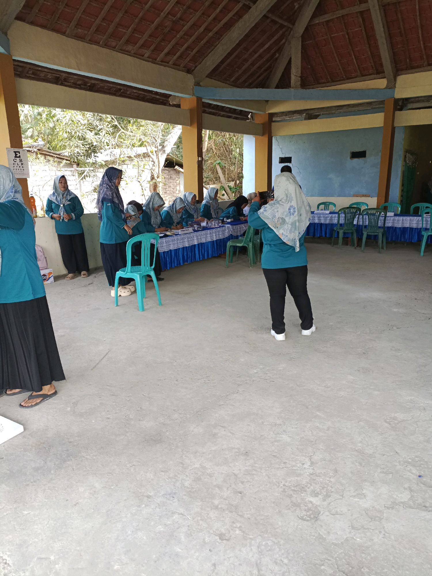 The image depicts an outdoor covered area, possibly a community hall or pavilion, in Rancang Kencono, Indonesia. A group of women, mostly wearing teal tops and headscarves, are gathered around tables set up for an event. Some are seated and appear to be engaged in administrative tasks, while others are standing. One woman in the foreground, facing away from the camera, is holding up a smartphone, seemingly taking a picture or recording a video. To the left, an eye chart is visible on a wall, suggesting a health-related activity. The environment is rustic, with a tiled roof supported by wooden beams and concrete flooring. Green plastic chairs are arranged around the tables. The overall impression is of a local gathering or registration event.
