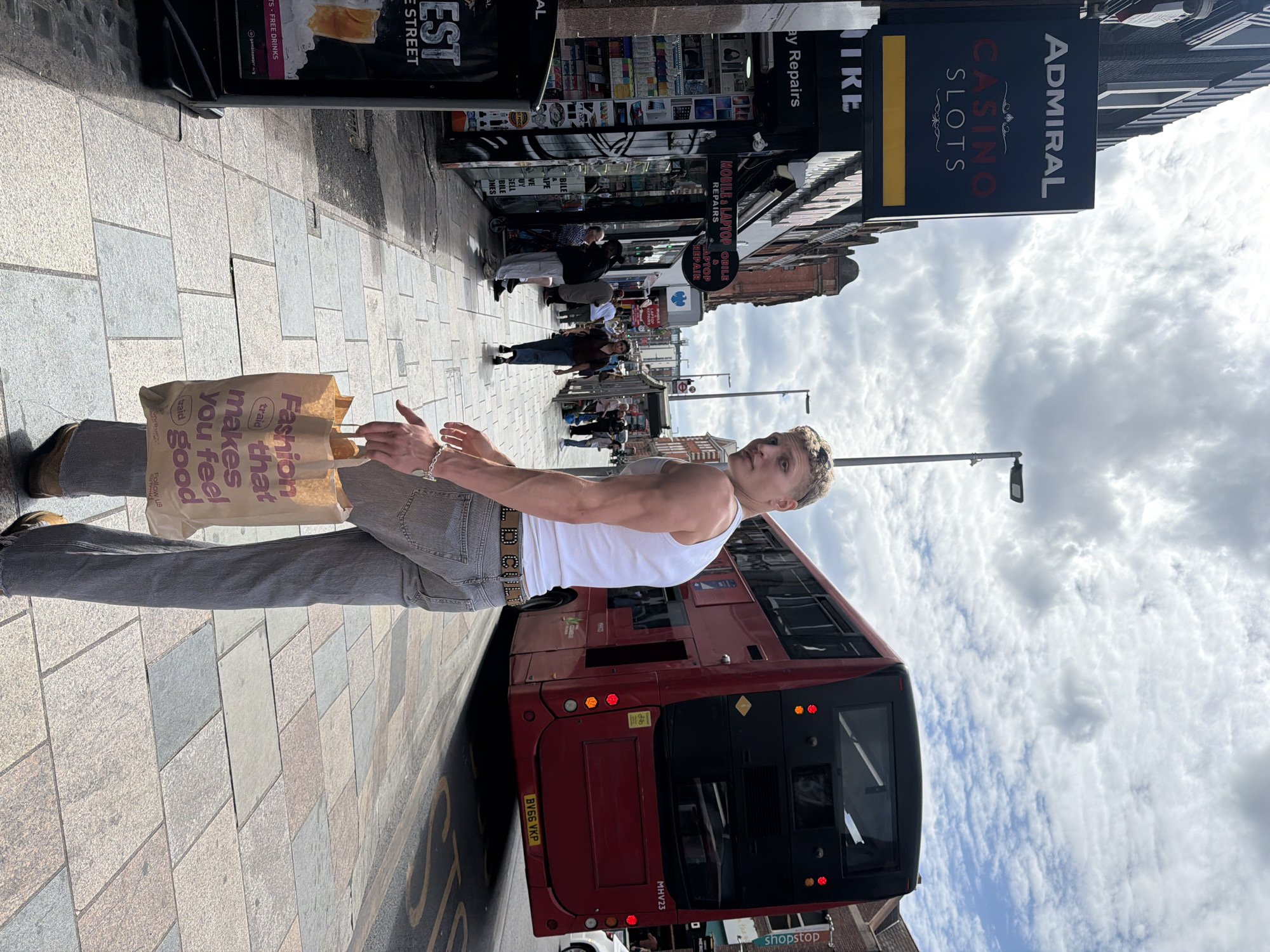 The image shows a young man standing on a sidewalk in what appears to be a busy commercial area of London, United Kingdom. He is wearing a white tank top, gray jeans with a studded belt, and carrying a shopping bag from "Fashion Traid." A red double-decker bus is positioned behind him, partially obstructing the view of buildings lining the street. The sidewalk is paved with patterned stones. Various storefronts are visible in the background, including "ADMIRAL CASINO SLOTS" and a mobile phone repair shop. The sky is cloudy. Several pedestrians are walking along the street. The bus has a license plate with the designation "BV66 VKP" and the bus number "MHV23".