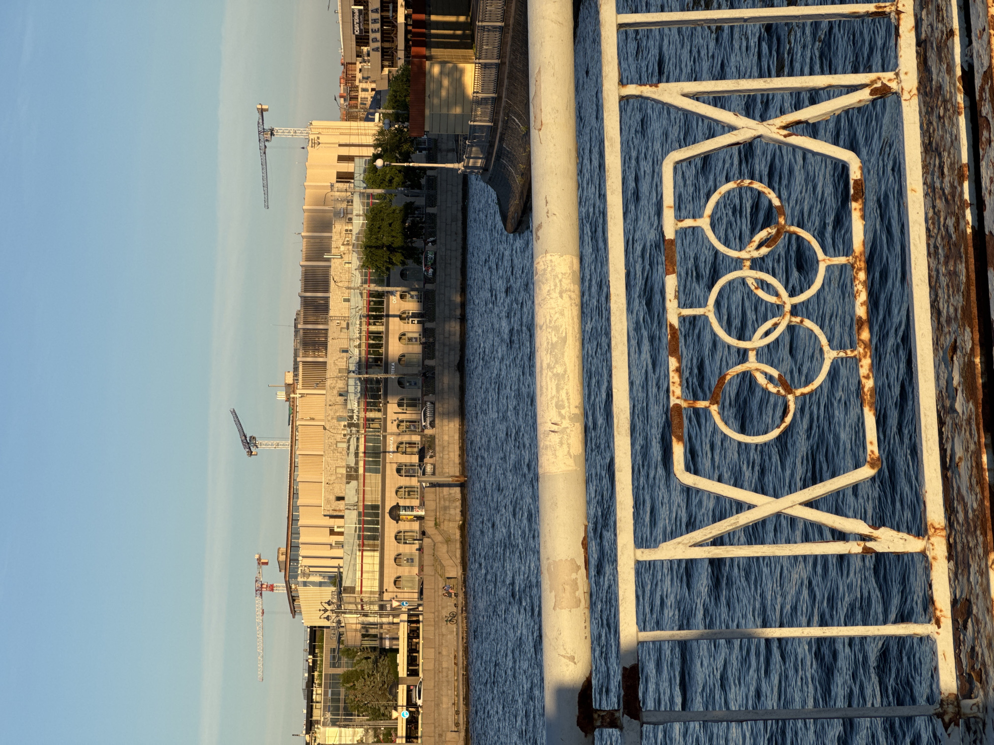 The image shows a close-up of a rusty white metal railing in the foreground, featuring a decorative pattern that resembles the Olympic rings, against a backdrop of the Neva River in Saint Petersburg, Russia. Beyond the river, a large, light-colored building complex with numerous windows and architectural details is visible. Construction cranes are present above the building, indicating ongoing development or renovation. The sky is clear and blue, suggesting a sunny day. The scene depicts an urban waterfront with elements of both established architecture and active construction.