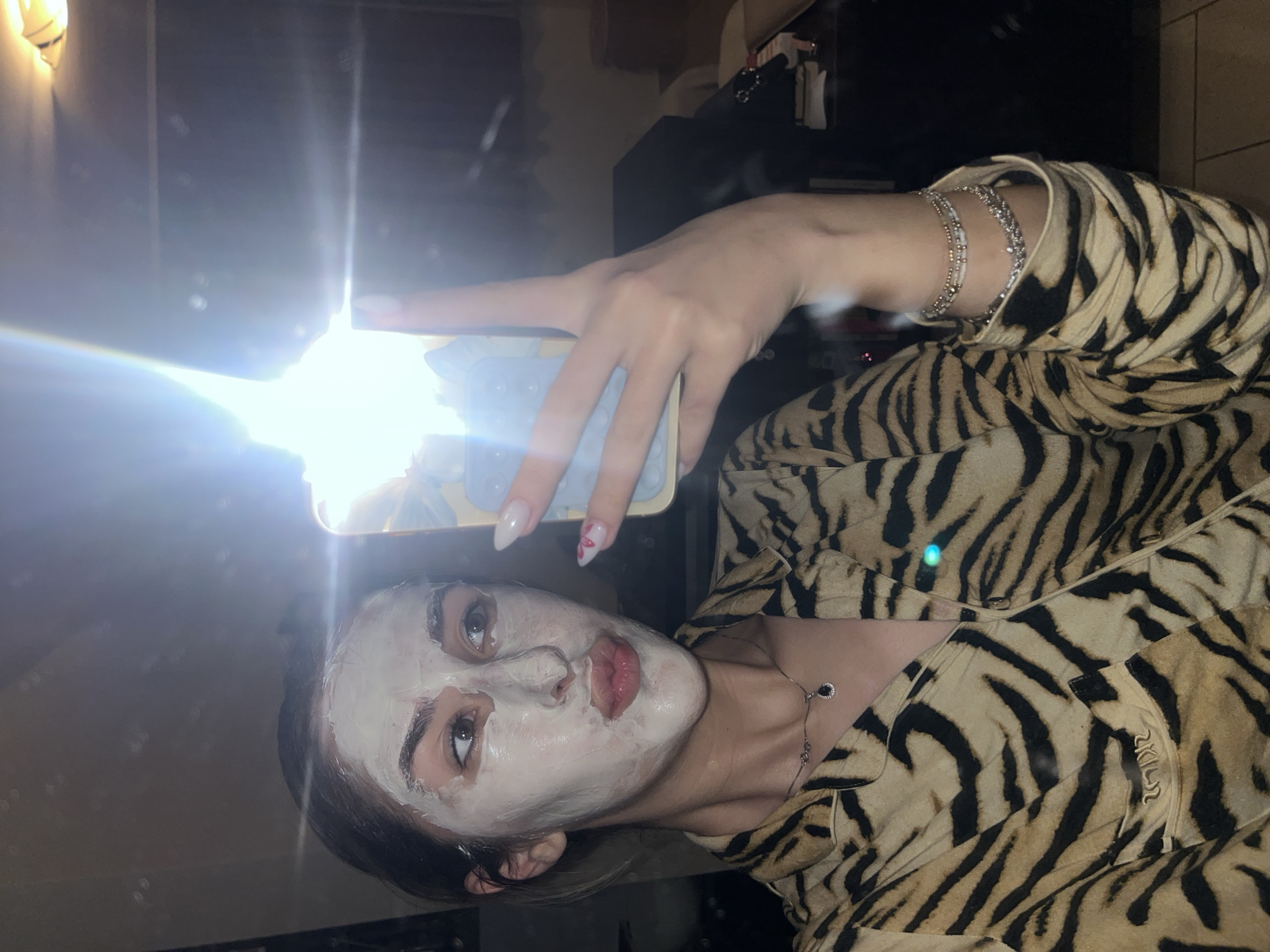 A person is holding a smartphone while looking into a mirror. The person is wearing a tiger-striped shirt and has a face mask on. The person also has several bracelets on their wrist and a necklace. The person's location is unknown.