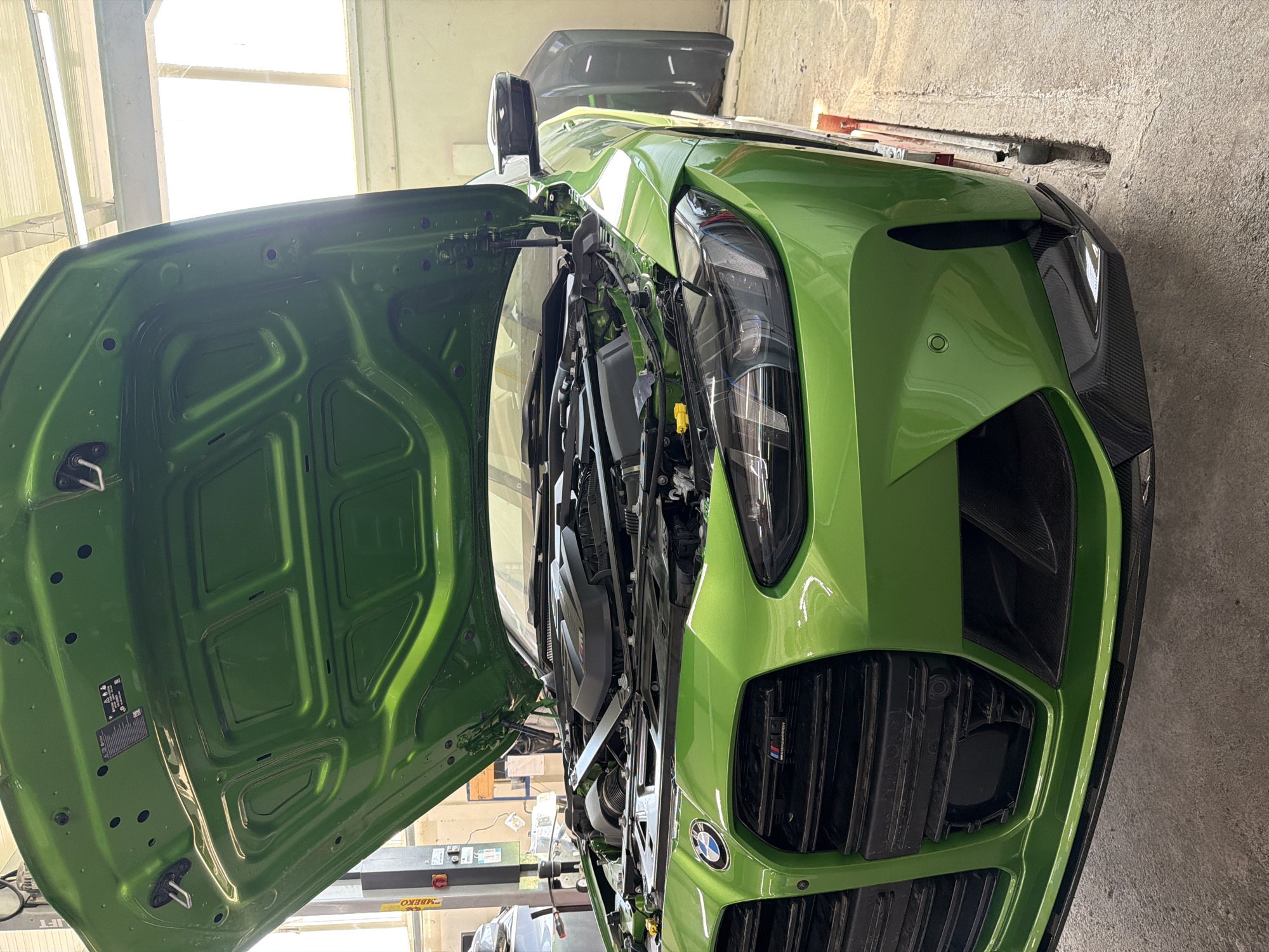 The image shows a lime green BMW with its hood open, revealing the engine bay. The car appears to be in a garage or workshop, possibly undergoing maintenance or repair. The location is not discernible from the image.