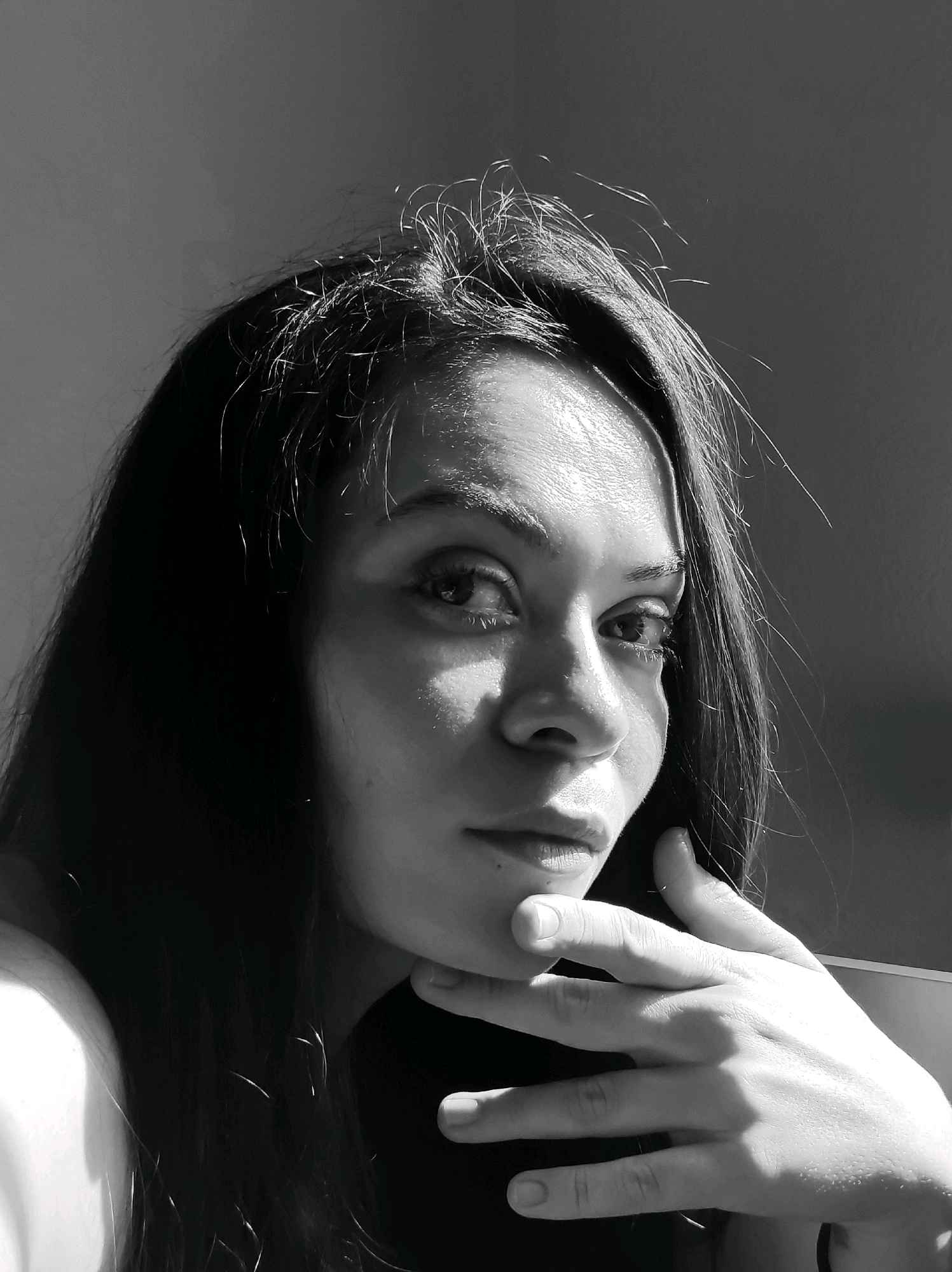 A black and white, close-up, waist-up studio portrait of a young woman with long, dark, wavy hair. She is positioned in the center of the frame and looking directly at the camera. Her right hand is raised, with her fingers gently resting on her chin and her thumb below her lower lip. The lighting creates strong highlights on her forehead, cheekbone, and nose, casting shadows across her face and hair. The background is a plain, dark, textured wall, with a subtle lighter tone on the left side of the frame. The overall mood is contemplative and dramatic due to the harsh lighting. The location is not discernible from the image.