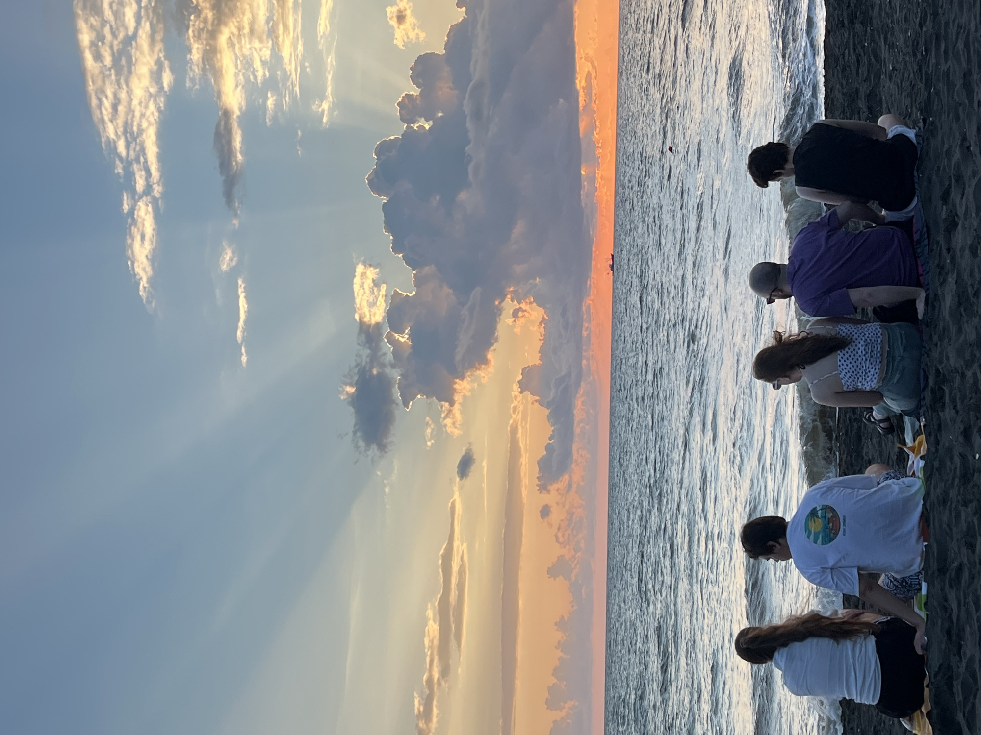 A group of five people are sitting on a dark sand beach in Ureki, Georgia, facing a body of water. The sky above is filled with dramatic clouds at sunset, with rays of light emanating from behind a large cloud formation. The water is choppy, with small waves rolling onto the shore. The people are dressed casually and appear to be watching the sunset. From left to right, the individuals are: a woman with long brown hair wearing a white shirt and black pants; a man with dark hair wearing a white t-shirt with a circular graphic; a woman with long brown hair wearing a white and black polka dot top and green shorts; a man with a shaved head wearing a purple t-shirt and white shorts; and a person with dark, curly hair wearing a black sleeveless shirt and white shorts. In the distance, a small boat is visible on the horizon. The scene captures a tranquil moment at dusk on the Black Sea coast.
