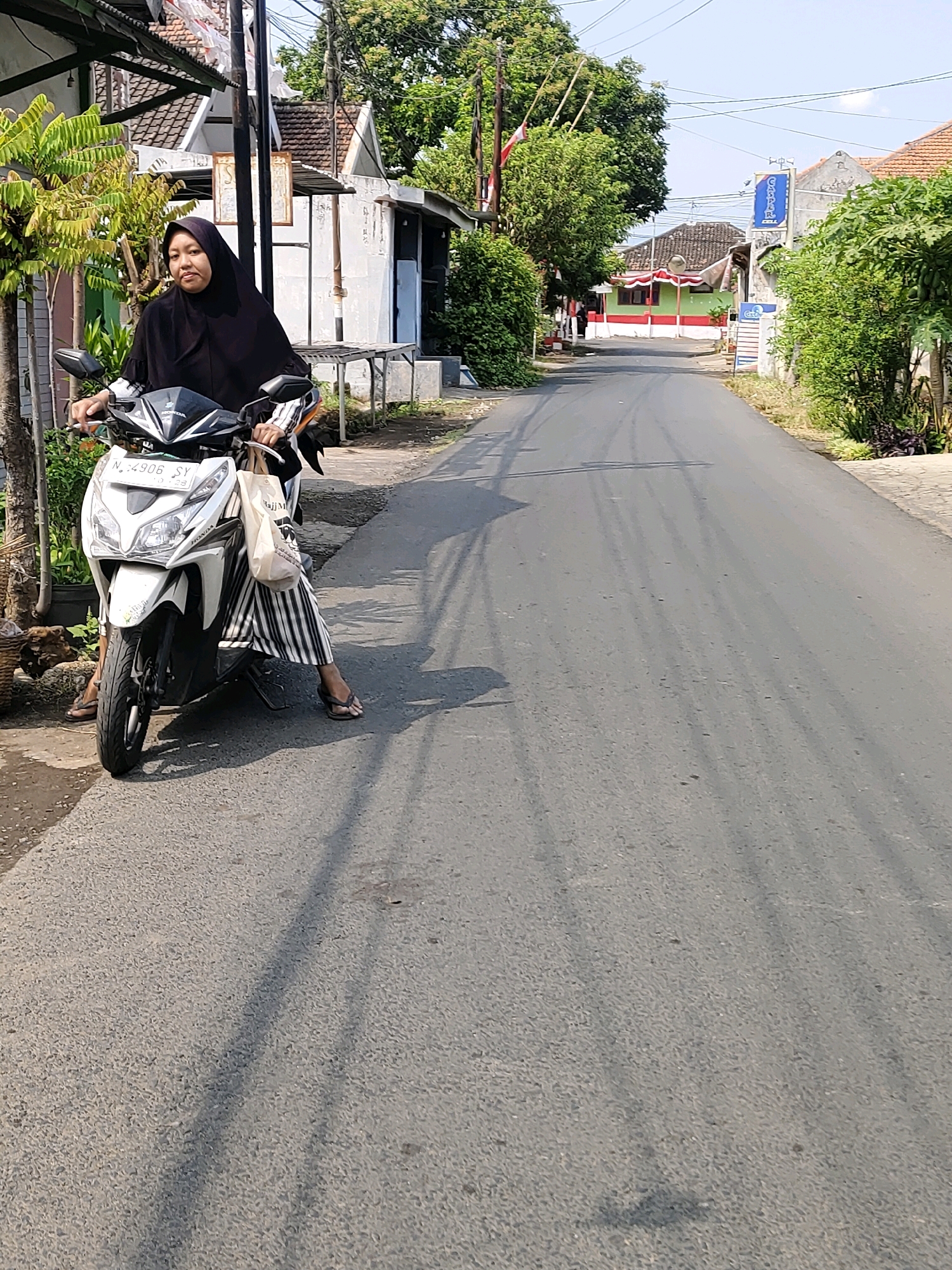 A medium shot captures a woman wearing a black hijab, seated on a white Honda Vario scooter parked on the side of a paved road. She is looking to her left, with a neutral expression. The scooter has a license plate that reads "N 4906 SY". She is holding a beige cloth bag in her right hand. The woman is wearing a black headscarf and a white and black striped skirt that reaches her ankles. Her feet are clad in brown flip-flops.
The environment is a residential street in Probolinggo, Indonesia, characterized by a mix of simple houses with tiled roofs and lush green trees. The road surface shows tire marks, indicating vehicular traffic. In the background, a red and white building with a green trim is visible, along with a blue sign that reads "GM PAR". The lighting suggests it is a sunny day, casting shadows from the trees and the scooter onto the road. The scene depicts a typical everyday moment on a street in an Indonesian city.