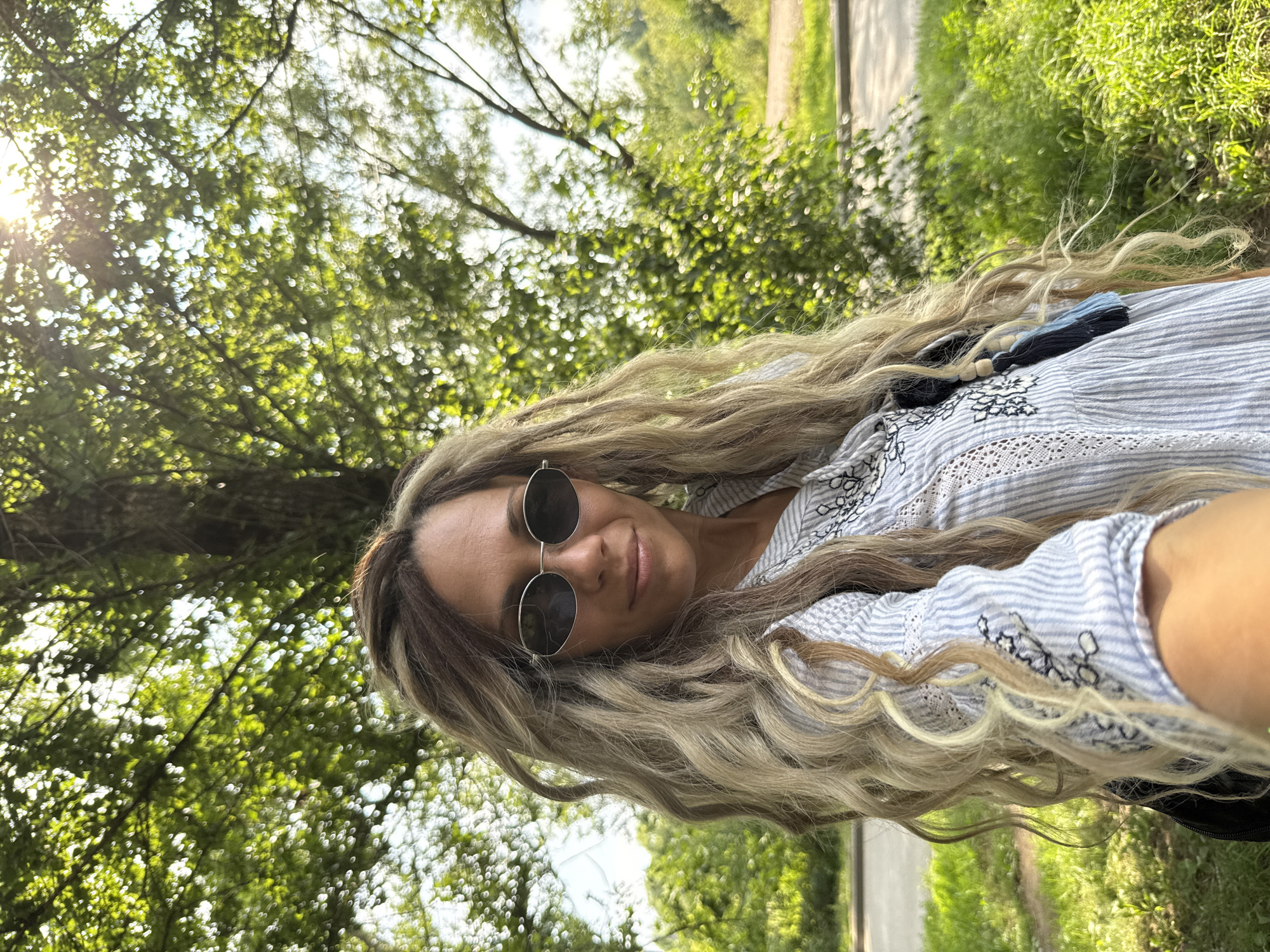 A woman with long, wavy, blonde and brown ombre hair, wearing oval sunglasses and a light blue and white striped shirt with embroidery and tassels, stands outdoors in what appears to be a park or wooded area. She is looking at the camera and smiling slightly. The background consists of green trees, foliage, and a dirt path or road. The sun is shining through the trees, creating a dappled light effect. The image suggests a casual, outdoor setting, possibly in or near Rusanovo, Russia, given the mention of this location. The woman is positioned in the foreground, with the natural environment filling the rest of the frame.
