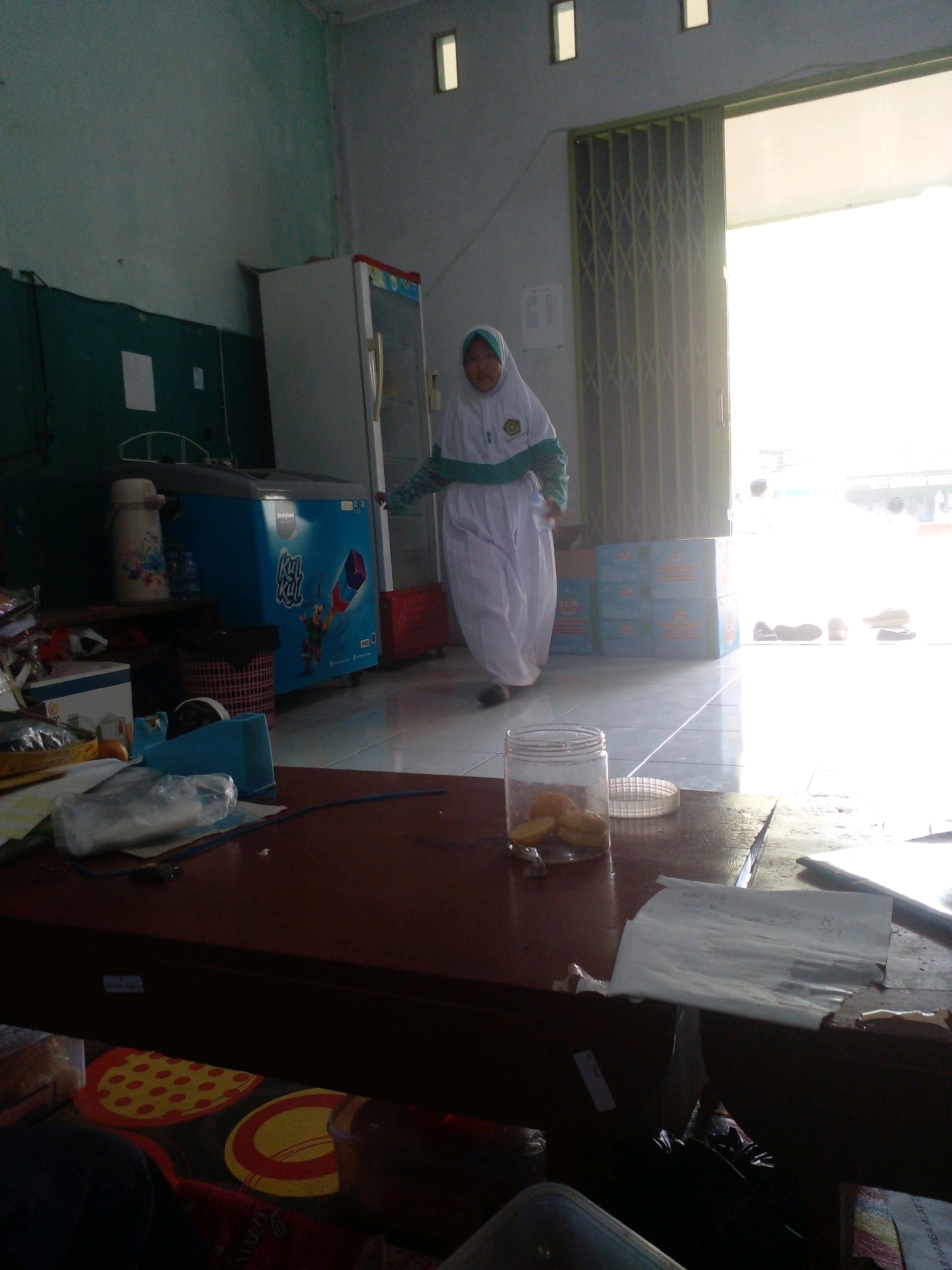 A person wearing a white hijab and white robe with teal accents walks through a room with a tiled floor. The room appears to be a shop or storage area. To the left, there is a blue and white refrigerator with graphics and a red stripe. Next to the refrigerator, on a shelf, are a beige thermos, a bottle of water, and a red basket. In front of the refrigerator, on a dark brown table, are various items including a clear plastic container with food, a roll of tape, and a plastic bag. To the right, sunlight streams in from an open doorway with accordion-style doors. Several pairs of shoes are visible on the floor near the doorway. The walls are painted a muted green and gray. The overall lighting is bright, especially near the doorway, indicating daylight. The location is identified as Banyuputih, Indonesia.