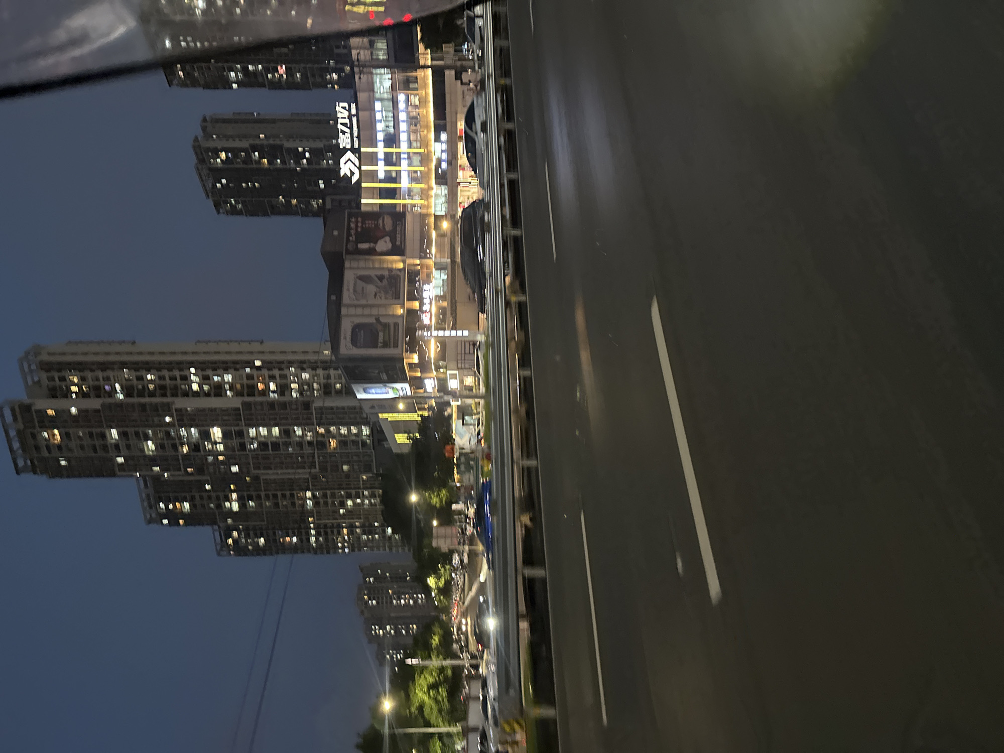 This is a night-time, wide-angle, eye-level shot of a busy urban environment, likely taken from a vehicle on a road. The foreground is dominated by a wet asphalt road with clear lane markings. Beyond the road, a metal guardrail separates the lanes of traffic from an elevated walkway or overpass. In the mid-ground, there is a steady stream of vehicles, with headlights and taillights creating streaks of light due to motion blur. Several tall, illuminated apartment buildings rise in the background, their windows glowing with warm light against the dark blue night sky. A commercial building with prominent signage, featuring Chinese characters, is visible between the apartment buildings. The overall impression is one of a bustling, modern city at night. This scene is located in the Chongqing High-tech Zone, China.