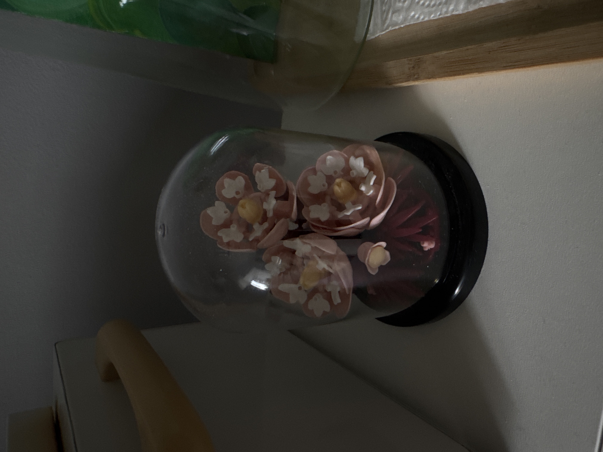 A close-up shot shows a glass cloche containing pink and white artificial flowers on a dark red base. The cloche has a black base. The object rests on a white surface next to a tan handle. Above the cloche is a wooden shelf with another glass object. The background consists of light grey walls. The location and information about Izhevsk, Russia, are not ascertainable from the provided image.
