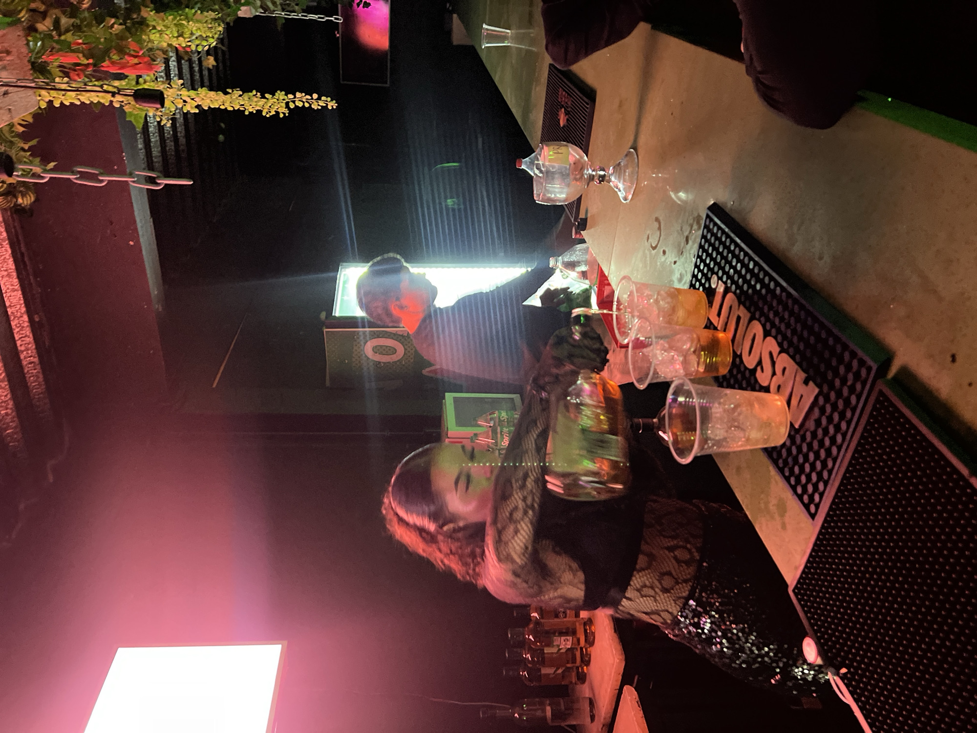 The image depicts a lively, low-lit bar scene at night in Arica, Chile, focusing on the activity behind a bar counter. The primary subjects are two individuals, likely bartenders or staff, and various bar equipment.
In the midground, a woman with brown, curly hair is actively preparing a drink, holding a clear bottle or shaker with both hands over the bar. She wears dark clothing and her face is illuminated by a green light. Behind her, several liquor bottles are arranged on a shelf. Another person, a man with short hair, is visible further back, illuminated by a bright, rectangular white light source. He appears to be looking towards the bar.
The bar counter, which dominates the right side of the frame, holds several clear plastic cups, some containing yellowish drinks with ice. A clear glass chalice with a small plastic bottle inside is also present. Two black rubber bar mats are on the counter, one of which clearly displays the brand name "ABSOLUT" in white text.
The setting is characterized by dim, artificial lighting, with strong pink/red and green glows creating a vibrant nightclub or bar atmosphere. In the background and top left, decorative elements include hanging artificial greenery and metal chains. The image captures the dynamic of drink preparation in a social, evening environment.