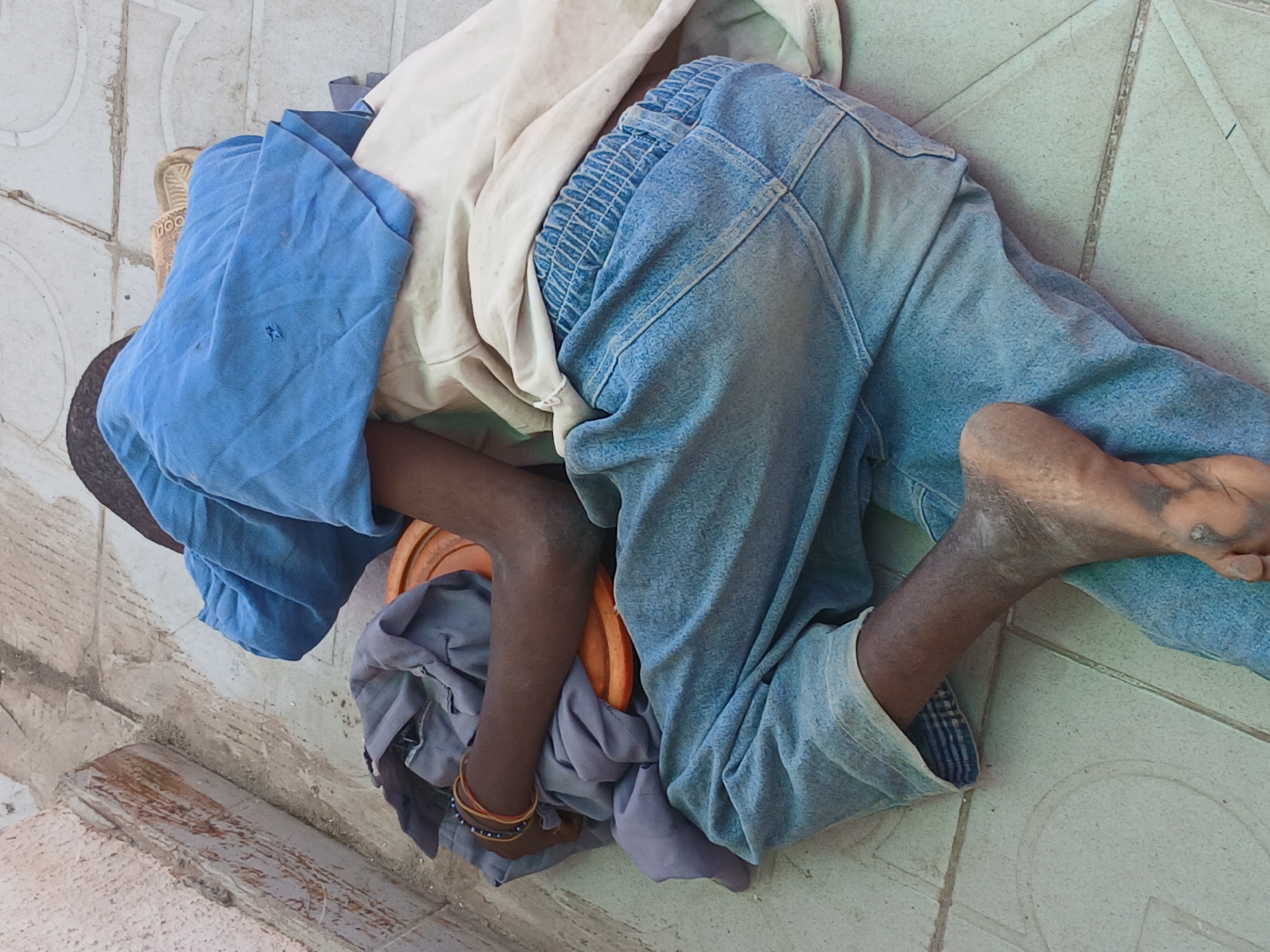 The image captures an overhead view of a person, likely a child or young individual, lying down on a light-colored tiled surface in Maiburiji, Nigeria. The person is positioned in a curled or fetal-like pose, with their face and upper body mostly obscured by a folded blue cloth, suggesting they are resting or sleeping.
They are dressed in simple, worn attire, including light blue denim jeans with an elasticated waist and a light-colored (possibly off-white) shirt. One arm is visible, adorned with a multi-colored beaded bracelet. Their bare feet and lower legs appear dusty and worn, indicating a lack of footwear.
Beneath the person's head/arm, a bundled grey cloth is visible, possibly serving as a makeshift pillow, alongside a partial orange object that resembles a plastic bowl or container. The tiled ground, featuring a geometric pattern, shows signs of dirt and wear. A textured, rough-edged concrete or wooden structure is partially visible in the bottom left corner of the frame. The scene portrays a moment of rest in what appears to be a public or outdoor setting, during daytime hours. There is no visible text in the image.