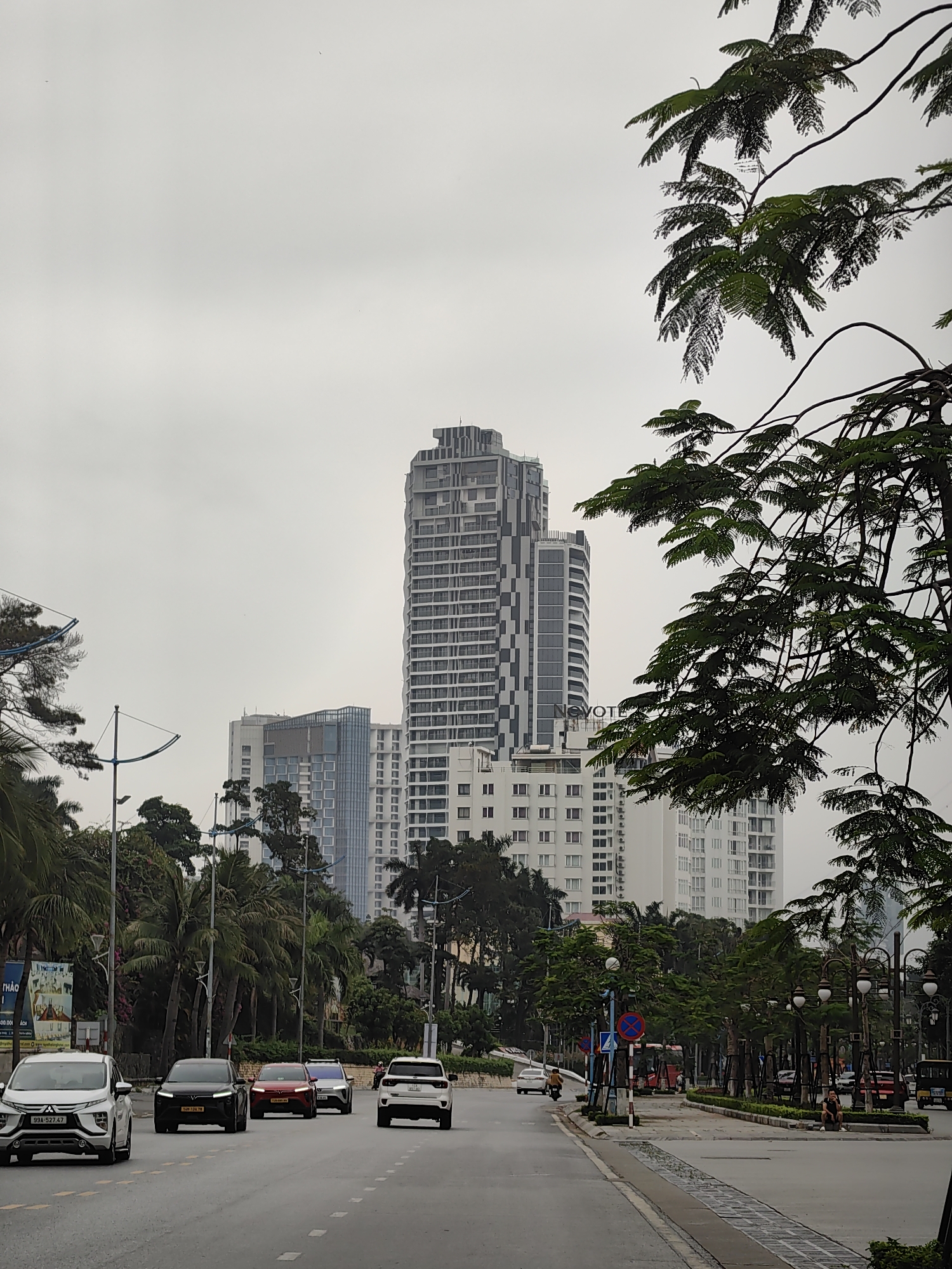 The photograph shows a street in Bai Chay Ward, Vietnam, with multiple buildings and trees. In the foreground, there are five vehicles driving down the road. There is a bus lane to the right of the photo with two pedestrians and various other vehicles. A tall building with a modern design is visible behind trees and smaller buildings. The sky is overcast and grey.