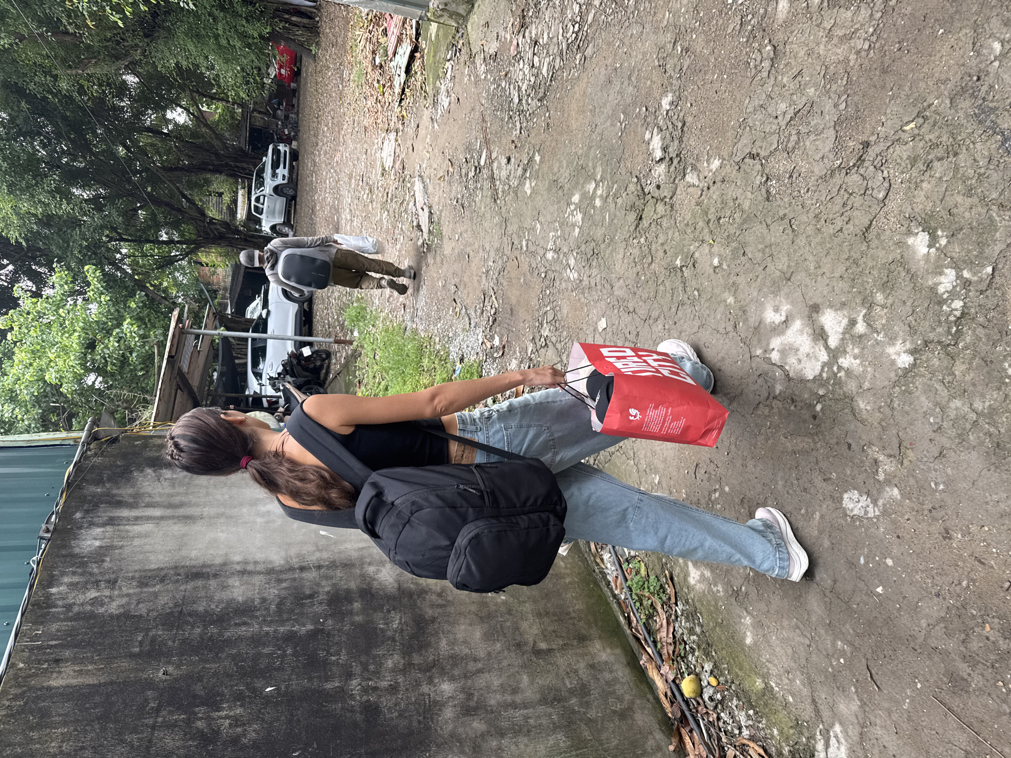 A young woman with dark hair in a low ponytail, wearing a black tank top and light blue jeans, is walking on a cracked concrete path. She carries a large black backpack and a red shopping bag. Another person walks ahead of her and wears a grey top with a backpack. There are parked cars on the side, and trees and green vegetation surround the path. The image was taken in Hà Nội, Vietnam.