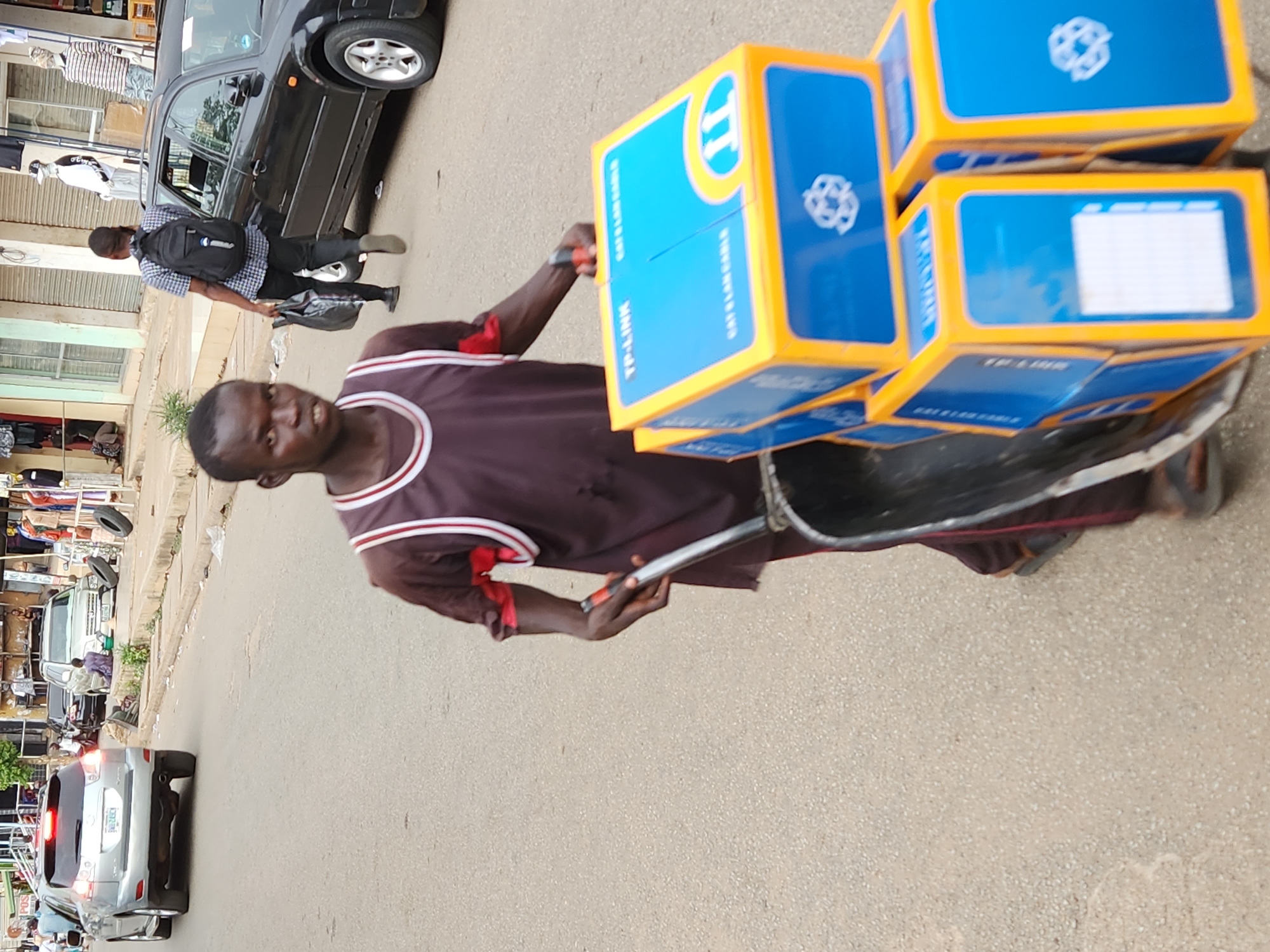 A man stands on a street in Jos, Nigeria, pulling a wheelbarrow piled with approximately six blue and yellow TP-Link CAT 6 LAN cable boxes. He is wearing a brown shirt with white and red trim. Another man with a black backpack walks past a black car in the background. Other vehicles and buildings are visible in the surrounding urban street scene.