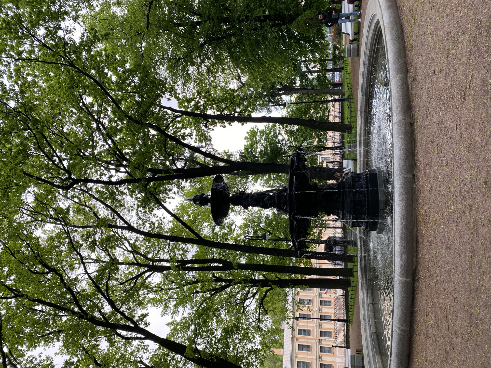 The image shows a fountain in a park, likely in Saint Petersburg, Russia, given the architectural style of the buildings visible in the background and the overall ambiance. The fountain is multi-tiered and dark in color, with water cascading into a circular basin. Trees surround the fountain, their branches reaching overhead and partially obscuring the sky. The ground surrounding the fountain is a mix of stone and dirt. In the background, a building with light-colored walls and multiple windows is visible behind a wrought-iron fence. Two people are standing near a bench, in the background. The day appears to be overcast or cloudy.