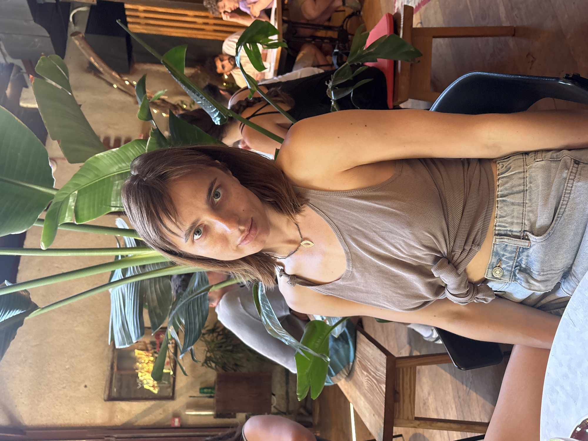 A young woman with brown hair, wearing a beige tank top and denim shorts, is seated at a table in a dimly lit, indoor establishment, possibly a cafe or restaurant. She is looking directly at the camera with a slight smile. Behind her, other patrons are visible, some seated at tables, and large green plants are prominent in the background, suggesting an indoor garden or a space with abundant greenery. The atmosphere appears casual and relaxed. The establishment's interior features exposed brick walls and wooden furniture, characteristic of a trendy urban setting. The presence of what appears to be a local Israeli brand on a bottle in the background, combined with the overall aesthetic, suggests the location could be Tel Aviv, Israel.