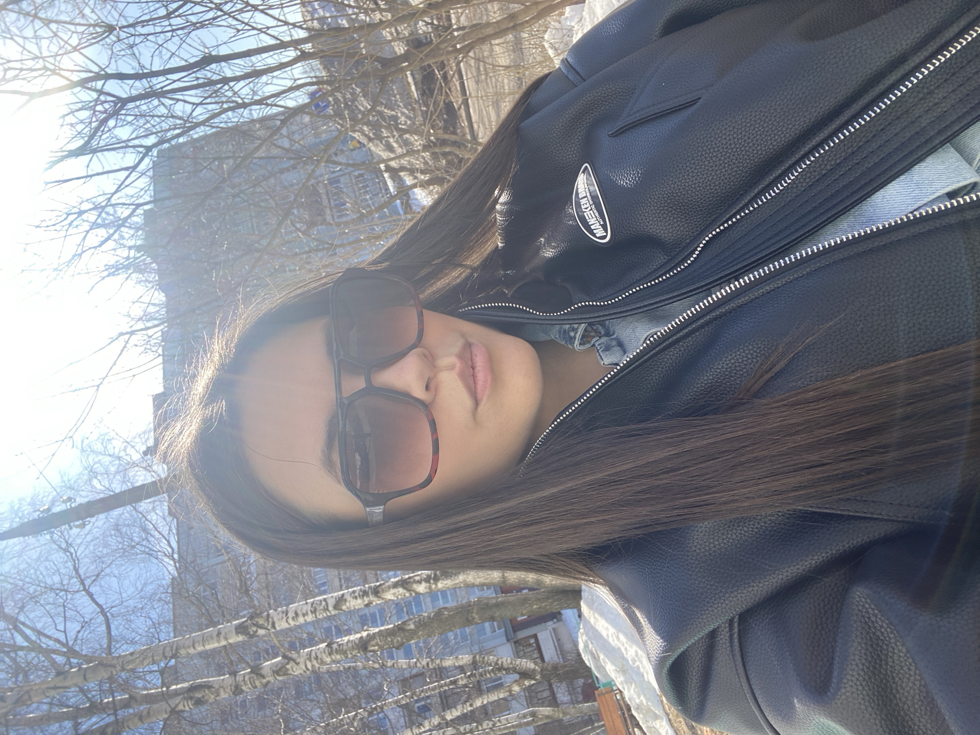 A young female is depicted in a close-up, chest-up self-portrait, centered in the frame. She has long, dark hair, which is illuminated by bright sunlight from the upper right, causing lens flare. She wears large, dark sunglasses with a visible tortoiseshell pattern on the frames, and her lips are slightly parted. Her attire consists of a black faux leather jacket, unzipped to reveal a light blue denim jacket underneath. A rectangular patch with the text "MANEKEN BRAND AUTHENTIC OVERSIZE APPAREL" is affixed to the right chest of the black jacket. The background indicates an outdoor urban environment, specifically a park or residential area in Chaikovsky, Russia. Patches of snow are present on the ground. Bare deciduous trees, including several distinct white-barked birches, populate the midground. A multi-story residential building is partially visible through the trees in the background. A brown wooden park bench is visible on the lower left. The scene suggests late winter or early spring conditions.