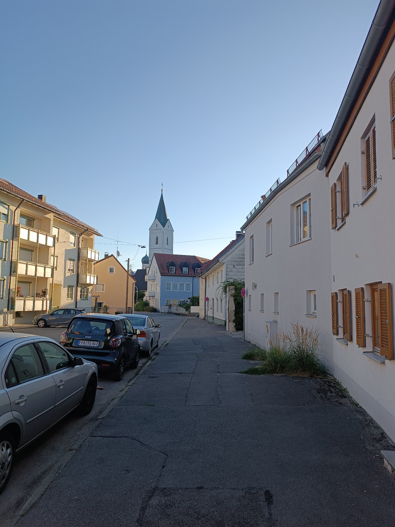This is a street-level, eye-level, medium shot of a street in the city of Fürstenfeldbruck, Germany, bathed in the warm light of a late afternoon or early morning sun. The scene is dominated by buildings on both sides of the street, a clear blue sky, and several parked cars.
On the left side of the street, a modern apartment building with cream-colored facades and balconies is visible. Its balconies are decorated with plants. Further down the street, a small, yellow-hued house stands, and beyond that, a pale blue building with a red-tiled roof. In the distance, a prominent white church tower with a green steeple rises above the other structures, identifiable as belonging to the parish church of St. Mary in Fürstenfeldbruck. The architectural style suggests a typical European town.
The right side of the street features a white building with wooden shutters on its windows. The building has a simple, clean facade. A patch of tall, dry grass grows in front of this building.
Several cars are parked along the curb on the left side of the street, facing away from the viewer. These include a silver sedan in the foreground, a black hatchback with a license plate reading "FFB TG 86E", and another silver car further back. A white car is visible parked on the right side of the street, partially obscured.
The street itself is paved with asphalt, showing some cracks and signs of wear. The sidewalk on the right is also visible. The overall impression is one of a quiet residential or mixed-use street in a German town. There are no people visible in the image. The lighting suggests a calm and serene atmosphere.