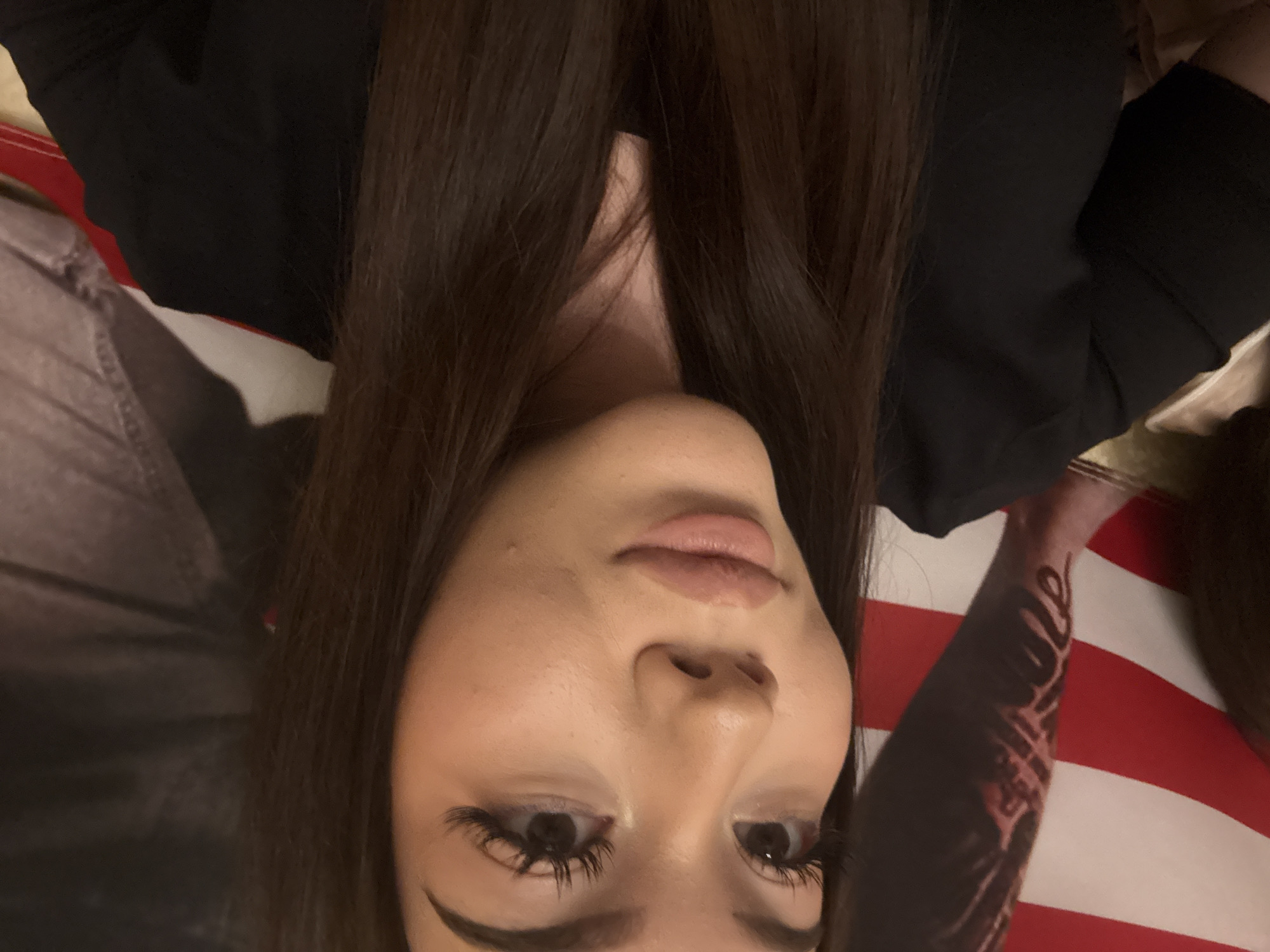 A close-up portrait shows a person, likely a woman, with long, dark brown hair flowing around her shoulders and chest. The image is taken from a slightly low angle, with the person looking downwards, giving a contemplative or introspective expression. Her face features clear skin, prominent winged eyeliner, and long false eyelashes, highlighting her eyes. Her lips are full and adorned with a light lip gloss. She is wearing a black top or garment.
On her right arm (visible on the left side of the image when upright), there is a distinct tattoo composed of script or lettering, which appears to be a stylized word, possibly "Love" or a similar term, though it's not fully legible.
The background primarily features a large American flag, identifiable by its prominent red and white stripes, partially obscured by the subject's arm and hair. The setting appears to be indoors, likely under artificial lighting, suggesting a personal space or a themed environment. The activity is a posed photograph, likely a selfie, capturing a moment of reflection. The presence of an American flag in Naberezhnye Chelny, Russia, is a notable contextual detail, suggesting a personal display or affinity.