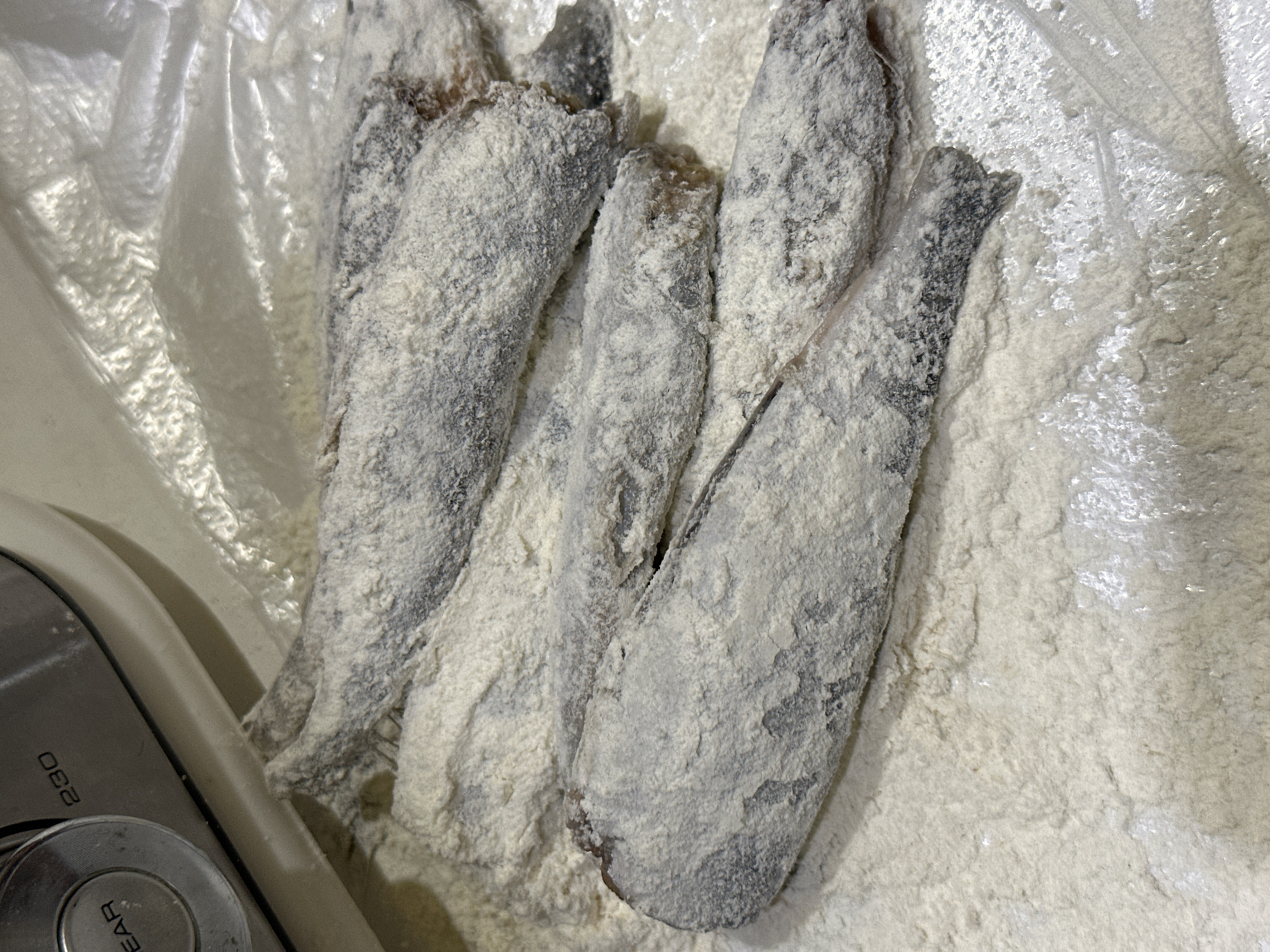 A close-up shot shows several raw fish, likely anchovies, that have been coated in flour and are lying on a surface of loose flour, possibly on a plastic sheet. The fish are dark with visible scales, and the flour adheres to them unevenly, creating a textured coating. In the upper left corner of the image, a portion of a kitchen appliance, possibly a food processor or mixer, is visible, featuring a silver dial with the number "230" imprinted on it. The lighting suggests an indoor setting, likely a kitchen.