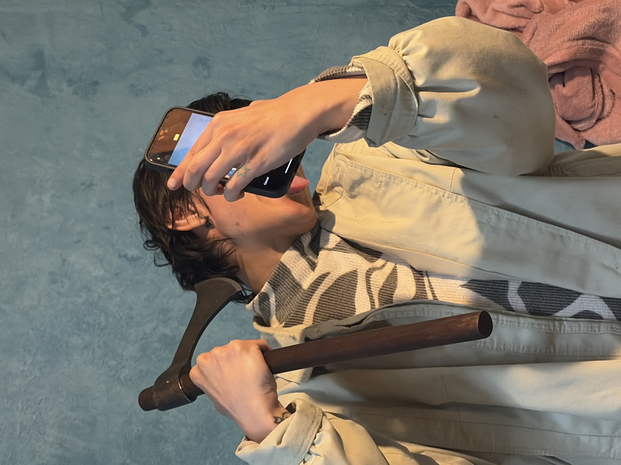 A person with dark hair is holding a dark-handled, single-bladed axe in their right hand, the axe head pointing left. They are simultaneously taking a selfie with a phone held in their left hand, sticking their tongue out. The person is wearing a beige jacket over a gray and white patterned top. The background is a pale blue wall on the left and a folded pink blanket on the right. No information in the image indicates the location is in Verinsko, Bulgaria.