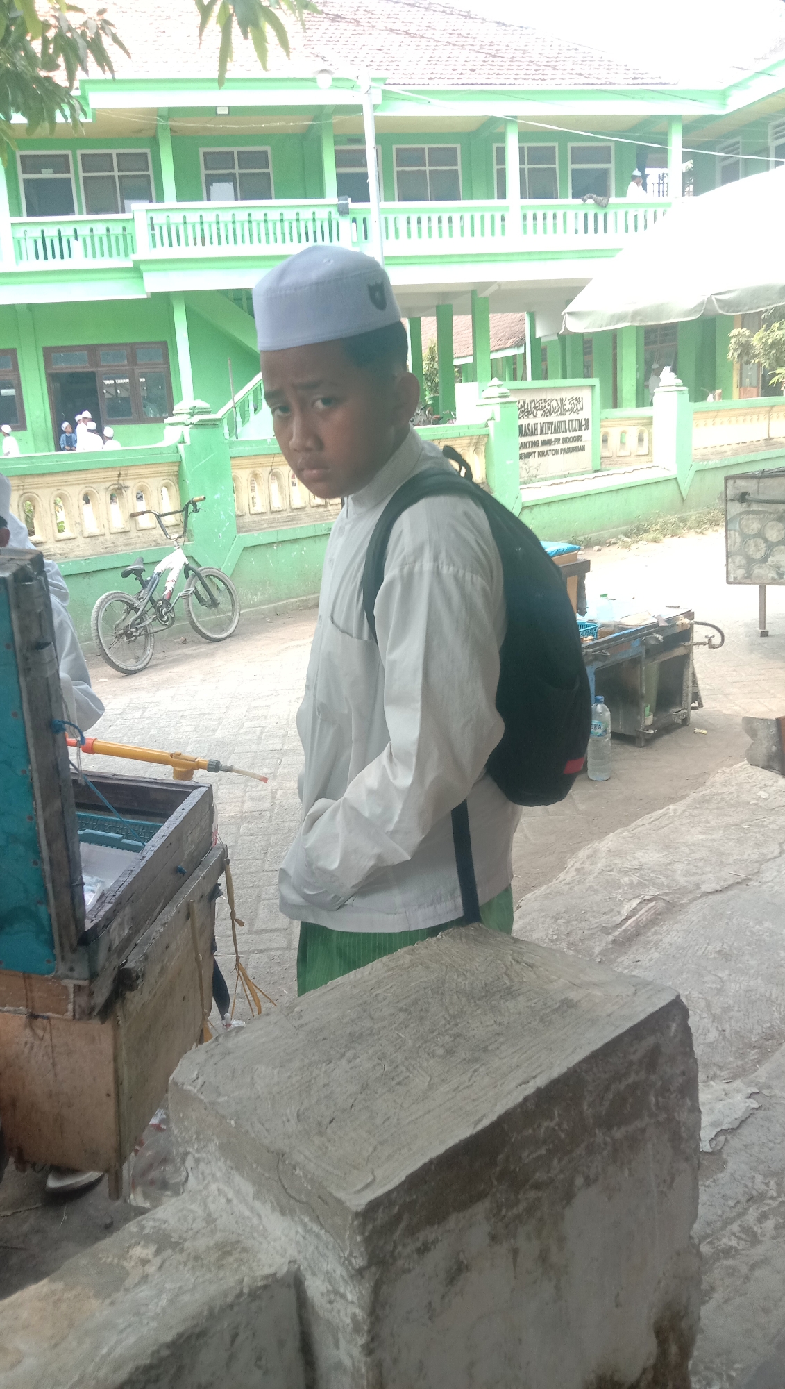 A young boy wearing a white kufi and a white long-sleeved shirt with a black backpack is standing in front of a green building with multiple levels. The building has a balcony with white railings and several windows. A sign on the building reads "MADRASAH MIFTAHUL ULUM-38" and has other text in Indonesian. To the left of the boy, a wooden cart with a blue top contains items. Behind the boy, a bicycle is parked. The ground is paved with concrete. The scene appears to be outdoors in a courtyard or school grounds in Ngempit, Indonesia, on a sunny day.