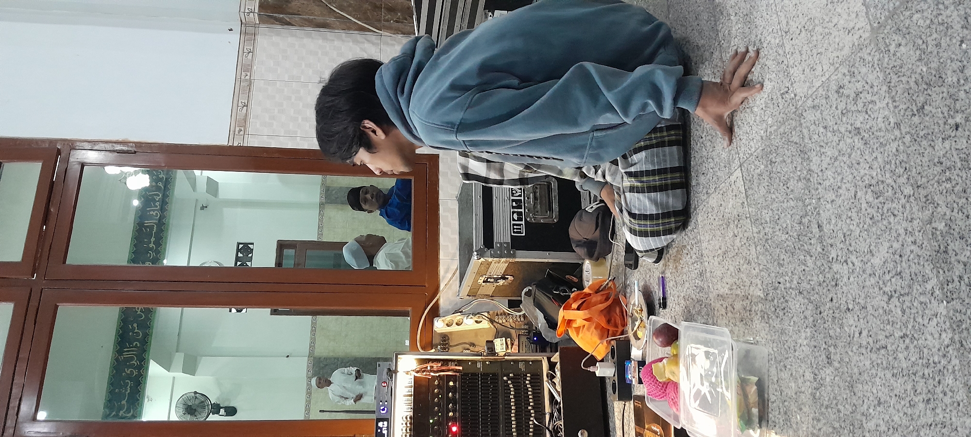 This image depicts an indoor scene, likely a mosque or community hall in Bendungan, Indonesia, given the presence of Arabic calligraphy on the walls and the attire of some individuals. In the foreground, a young person is seated on the floor, wearing a blue hoodie and checkered pants, looking towards a reflection in a large window. The reflection reveals three men. One man, dressed in white with a skullcap, stands holding a microphone. To his right, two other men are seated, one in a white skullcap and the other in a blue shirt.
The environment is cluttered with audio equipment, including a large mixing console with numerous knobs and lights, several black flight cases, and various cables. An orange bag, a roll of tape, and a power strip with multiple outlets are also visible on the floor. In the lower left corner, a clear plastic container holds several pieces of fruit, including what appears to be a pink pomelo, an apple, and other unidentifiable items. The floor is tiled with a grey and black speckled pattern. The overall impression is one of preparation for an event, possibly a religious gathering or a live performance, within a space that also serves as a place of worship.