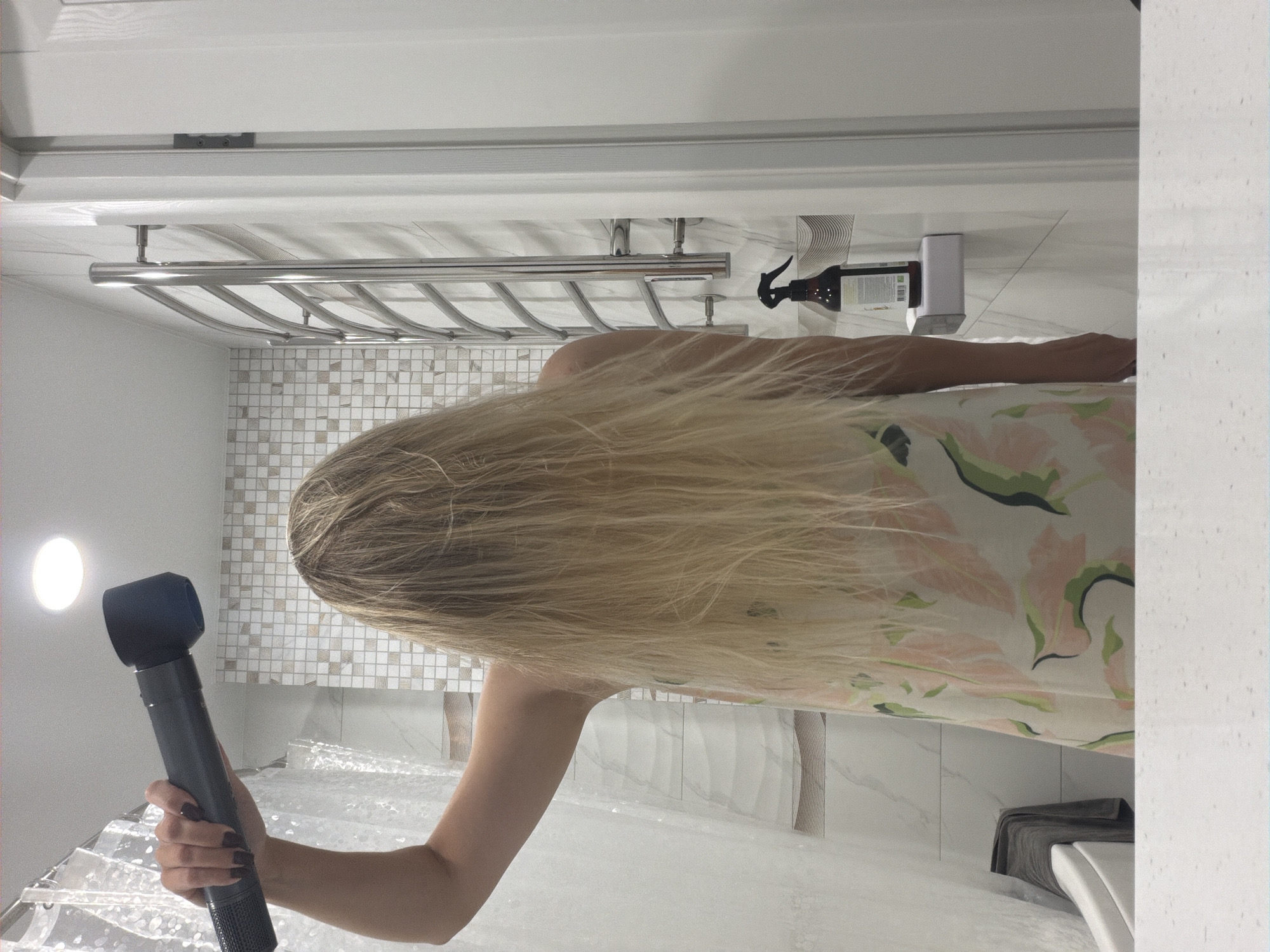 A person is in a bathroom, holding a hairdryer to their wet blonde hair. The person is wearing a floral-patterned robe or dress. The bathroom features a tiled wall, a towel rack, and a shower curtain. A bottle of product is visible on a shelf. The image was taken in Votkinsk, Russia.
