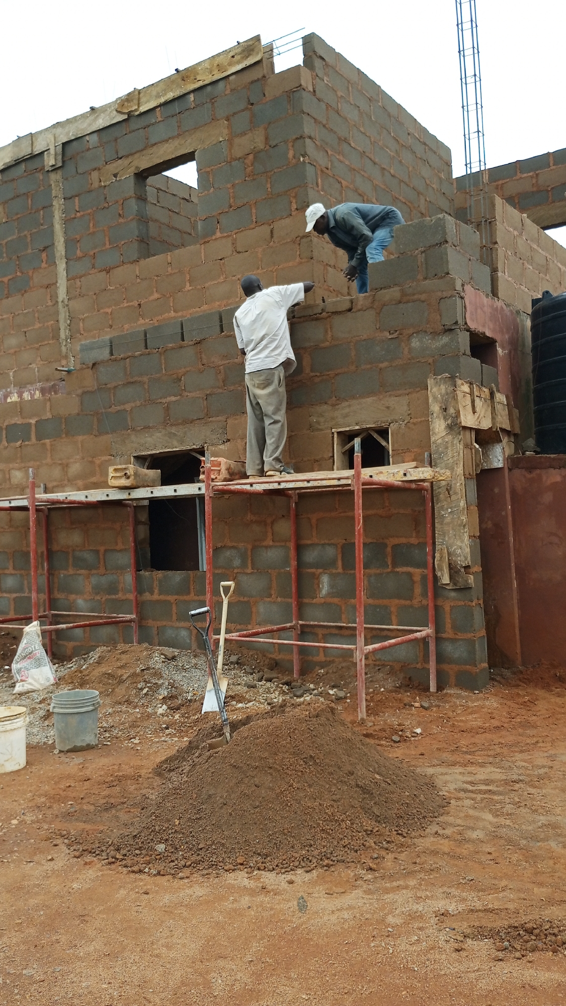 In Kuru Dungus, Nigeria, two construction workers are observed on a building site. The scene depicts the ongoing construction of a brick building. One worker, dressed in a white shirt and grey pants, stands on scaffolding, facing away from the camera and appearing to be working on the wall. The second worker, wearing a dark jacket, white cap, and blue jeans, is positioned on a higher level of the structure, leaning over the wall and also seemingly engaged in construction.
The building itself is constructed from cinder blocks, with visible wooden planks and rebar indicating the early stages of structural development. Scaffolding made of red metal pipes supports a wooden plank platform where the first worker stands. Tools, including a shovel and a trowel, are visible on the ground and on the scaffolding. A large pile of dirt or sand sits in the foreground, alongside two buckets and a bag of cement, suggesting active construction work. A black water tank is visible on the right side of the image, attached to the building. The ground is covered in reddish-brown soil, characteristic of the Nigerian terrain. The sky is overcast.