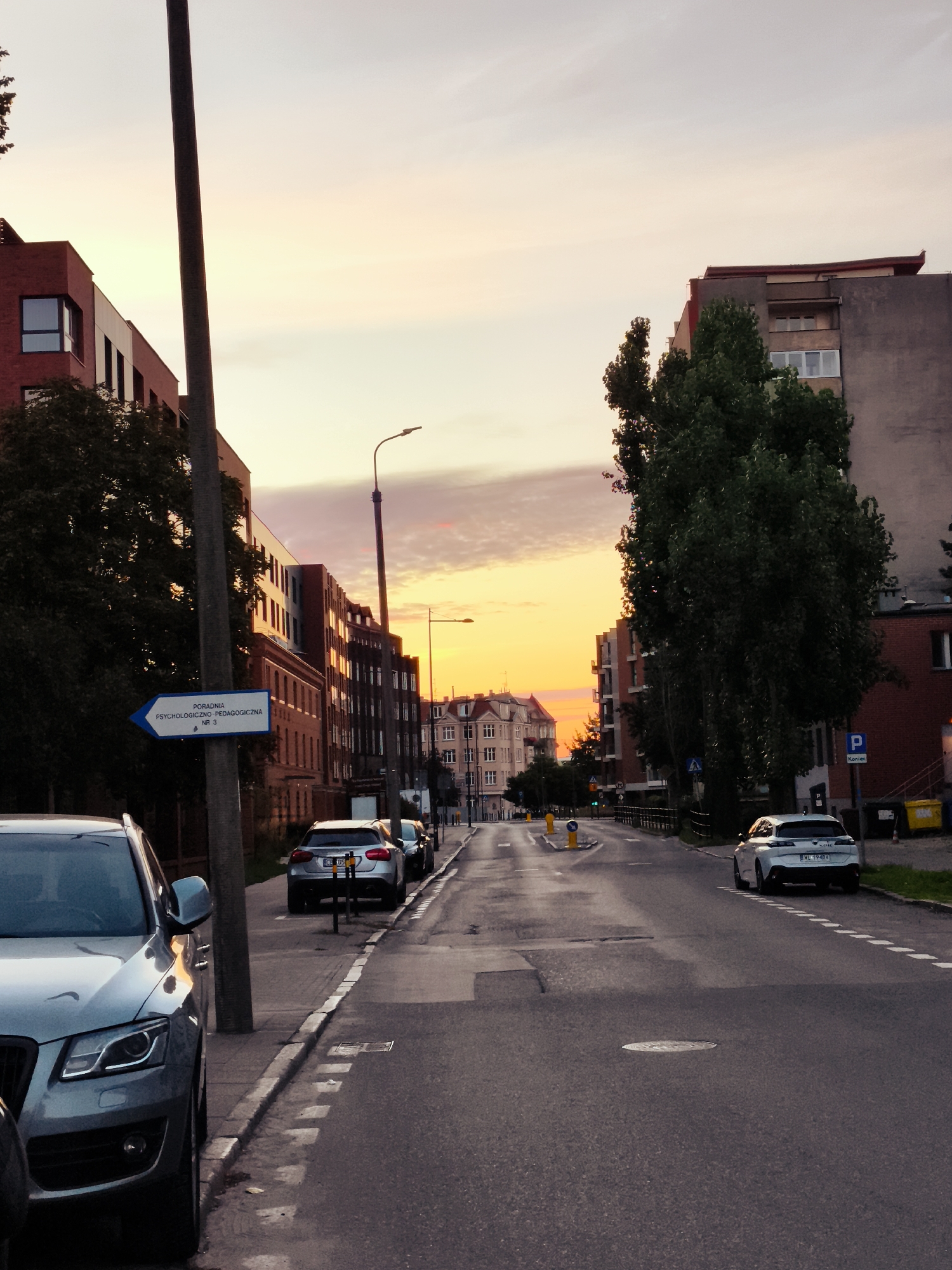 The image depicts a street scene in the city of Gdańsk, Poland, during sunset. The sky displays a gradient of warm colors, transitioning from yellow to pink. On the left side of the street, a modern brick building with dark windows is visible, adjacent to a taller, older-looking building. A tall lamppost stands prominently in the foreground, with a blue directional sign attached to it indicating "PORADNIA PSYCHOLOGICZNO-PEDAGOGICZNA NR 3" (Psychological-Pedagogical Clinic No. 3). Several cars are parked along the street, including a silver SUV in the foreground on the left and a white hatchback on the right. The street itself is asphalt, with painted white lines marking parking spaces and the lane division. In the background, more buildings and trees line the street, suggesting an urban environment. The lighting suggests it is either early morning or late evening.