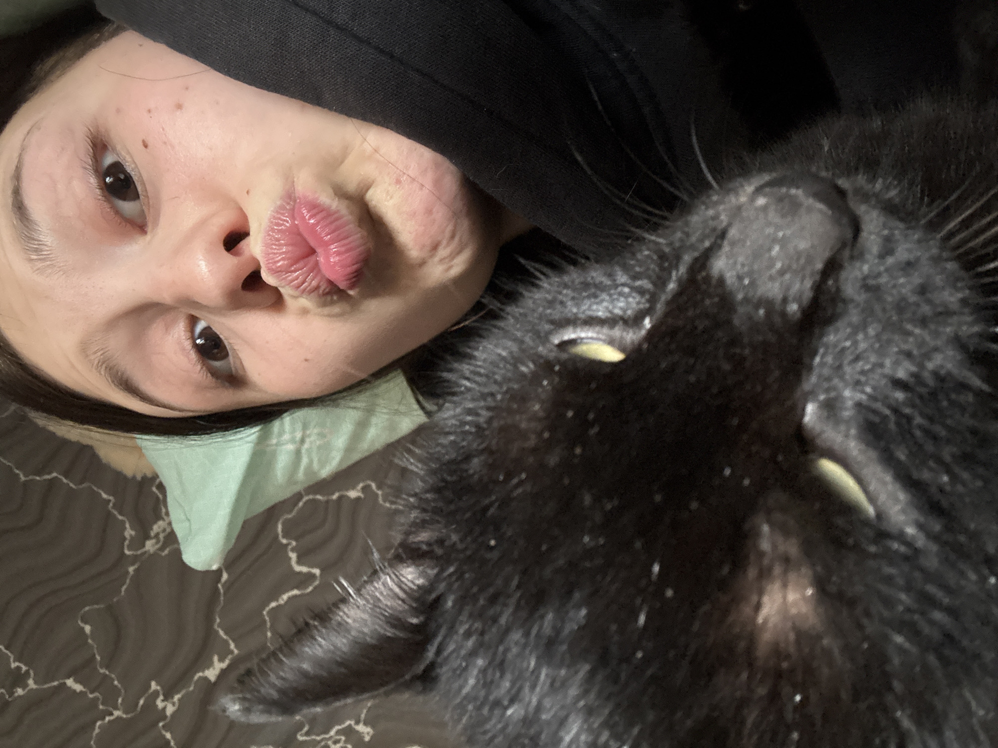 A selfie shows a person with fair skin and pouting lips lying next to a black cat. The person's eyes are brown, and their hair is dark. They are wearing a black hoodie. The cat is large and fills much of the right side of the frame. It has yellow eyes and a glossy coat. They are lying on a surface with a mottled, marbled pattern in dark gray and white. Part of a light green fabric, possibly a pillow or shirt, is visible beneath the person's head. There is no indication of the city of Фоки, Russia in this image.