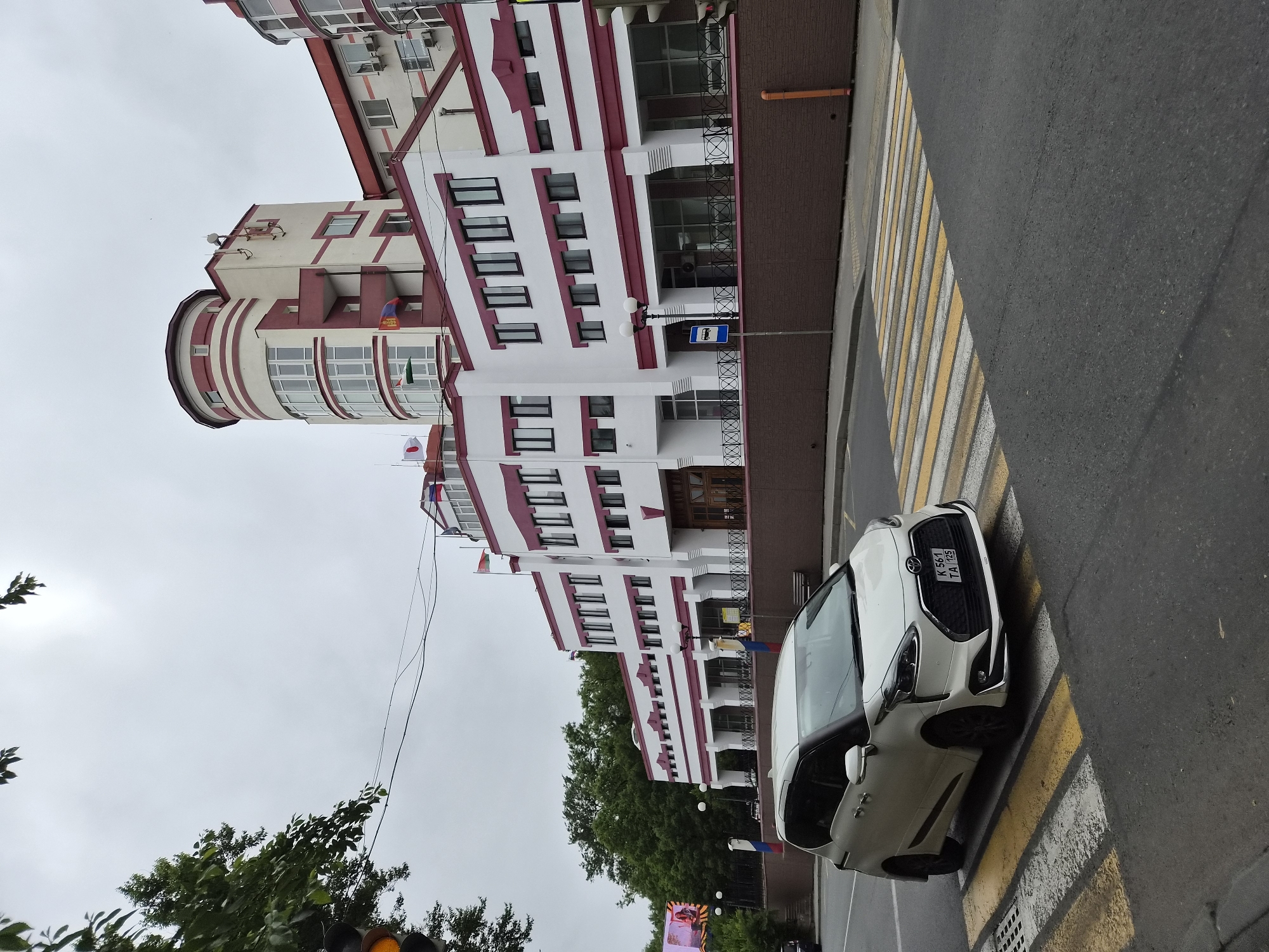 The image shows a white minivan stopped at a pedestrian crossing in front of a modern building in Vladivostok, Russia. The building is white with maroon accents and features a distinctive cylindrical tower section. Flags are visible on the building's facade. The sky is overcast. The minivan has a license plate that reads "K 561 TA 125". The pedestrian crossing is marked with yellow and white stripes. Trees and traffic signals are also visible in the background, indicating an urban environment.
