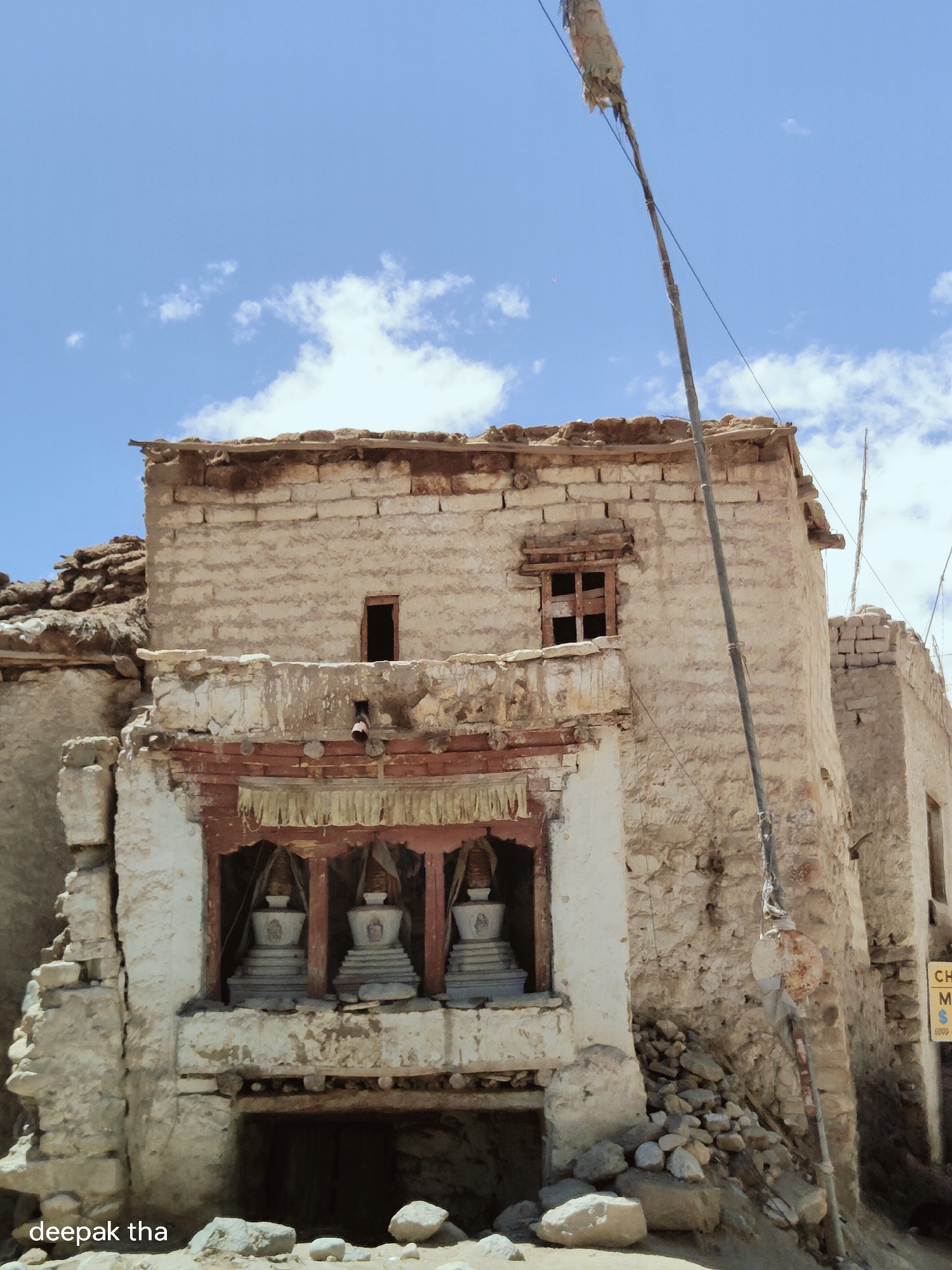 The image shows an ancient, weathered Buddhist monastery in Leh, India. The structure, built from mud bricks and stone, exhibits signs of decay and age. In the central facade, three white stupas are visible in arched niches, each topped with a spire and umbrella-like decoration. These stupas are framed by a wooden structure with decorative fabric hangings above. The surrounding buildings appear to be in a similar state of disrepair, with rubble and loose stones scattered around the base. A tall, thin pole with a tattered flag or cloth attached to it stands to the right of the main structure, extending upwards towards a bright blue sky with scattered white clouds. The overall scene conveys a sense of history and the enduring presence of Buddhist heritage in the high-altitude region of Leh.