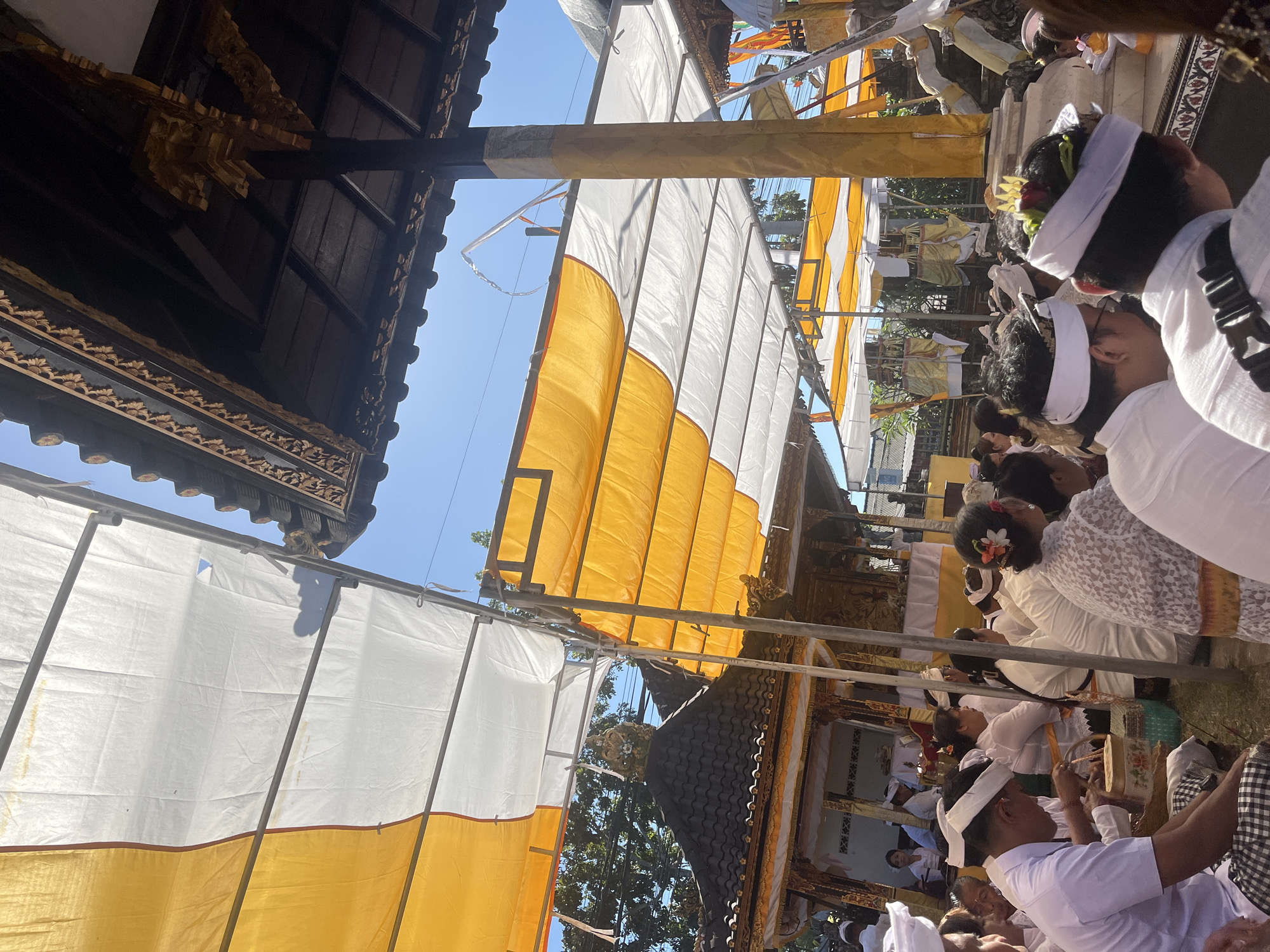 In Mendoyo, Indonesia, a ceremony is taking place under a canopy of yellow and white fabric. The structure, possibly a temple or a dedicated ceremonial space, features traditional Balinese architecture with dark wood and ornate golden carvings. A large yellow pillar supports the canopy, adorned with decorative fabric. Numerous people are gathered, many wearing white or light-colored traditional attire and head coverings, suggesting participation in a religious or cultural event. Some individuals are seated, while others appear to be observing or engaged in ritualistic activities. The scene is bathed in sunlight, indicating an outdoor setting under a clear blue sky. The presence of offerings or decorative elements within the building suggests a sacred or celebratory occasion.