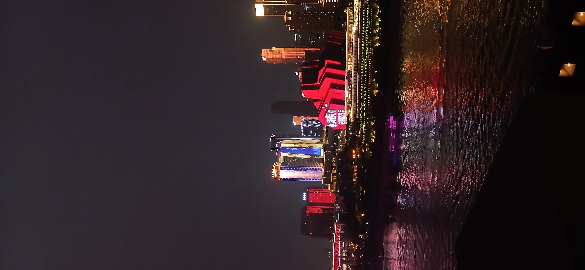The image shows a nighttime view of the city of Nan'an District, China, with the skyline illuminated by colorful lights. A river runs along the right side of the frame, reflecting the city lights. The buildings display various shades of red, blue, and purple. Some buildings have bright displays, including text in Chinese characters. The sky is dark, and the water is rippled.
