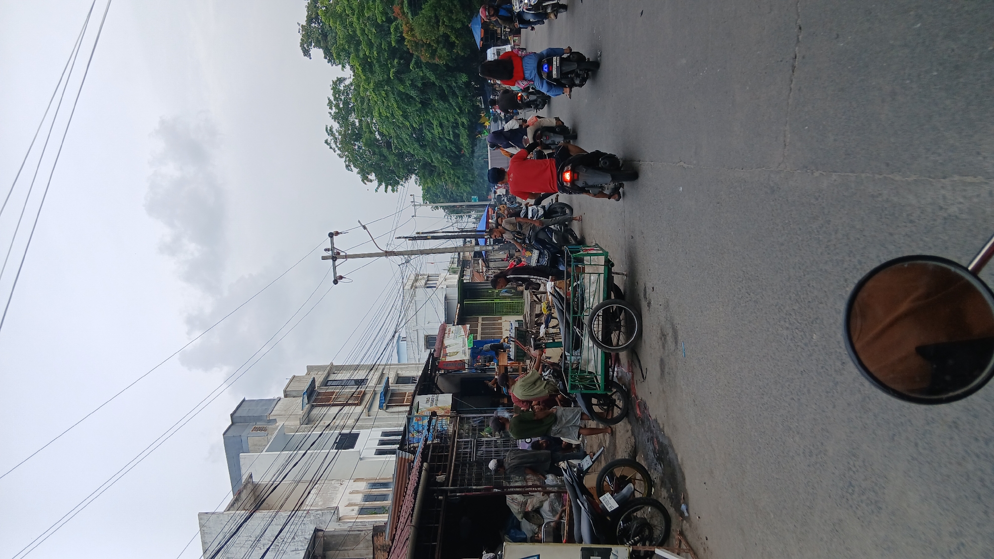 The image depicts a street scene in the City of Medan, Indonesia, likely a commercial area with buildings lining one side of the road and a line of trees on the other. The sky is overcast with a pale gray hue. The street is paved and appears to be moderately busy with motorcycles, some carrying riders and others parked. On the left side of the street, there are several buildings with storefronts. Some appear to be simple structures with corrugated metal roofs, while others are multi-story buildings with visible windows. There are several people visible, mostly men, standing and interacting near the storefronts. One person is riding a motorcycle with a green trailer attached. Further down the street, more motorcycles are in motion, creating a sense of traffic flow. The overall atmosphere suggests an active marketplace or a bustling urban thoroughfare.